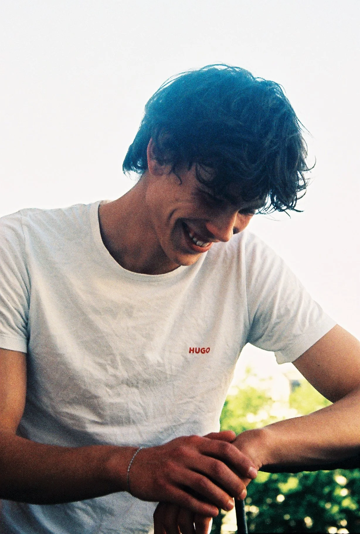 Young man with curly dark hair wearing a white Hugo t-shirt, smiling and looking down outdoors during daytime.