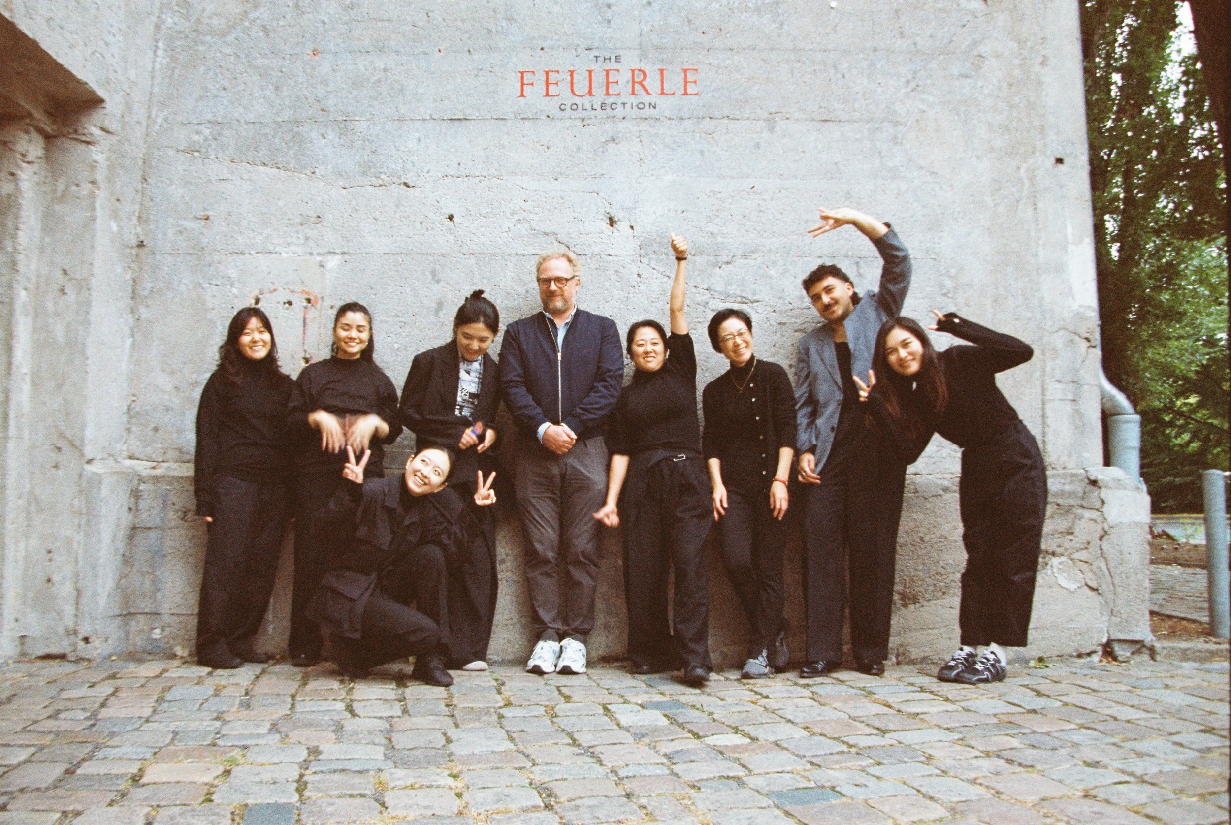 Group of eight people posing in front of a concrete wall with the words 'The Feuerle Collection' painted on it, some making peace signs and smiling.