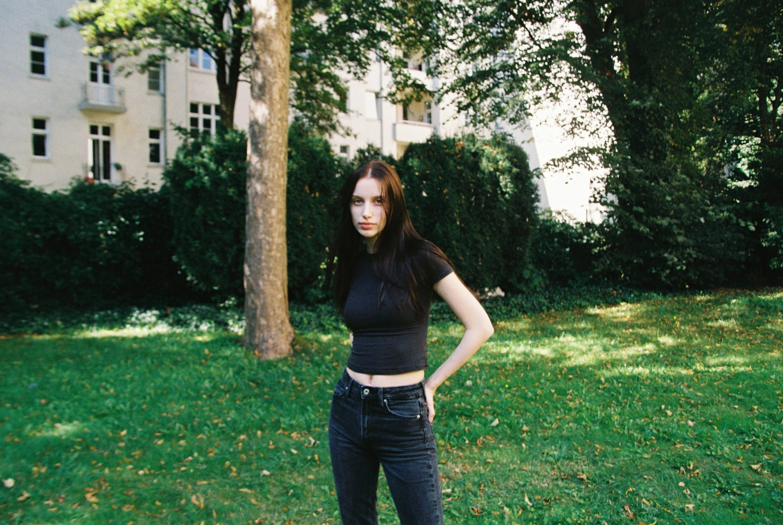 A young woman with long dark hair wearing a black t-shirt and black jeans standing outdoors on a grassy area with trees and apartment buildings in the background.