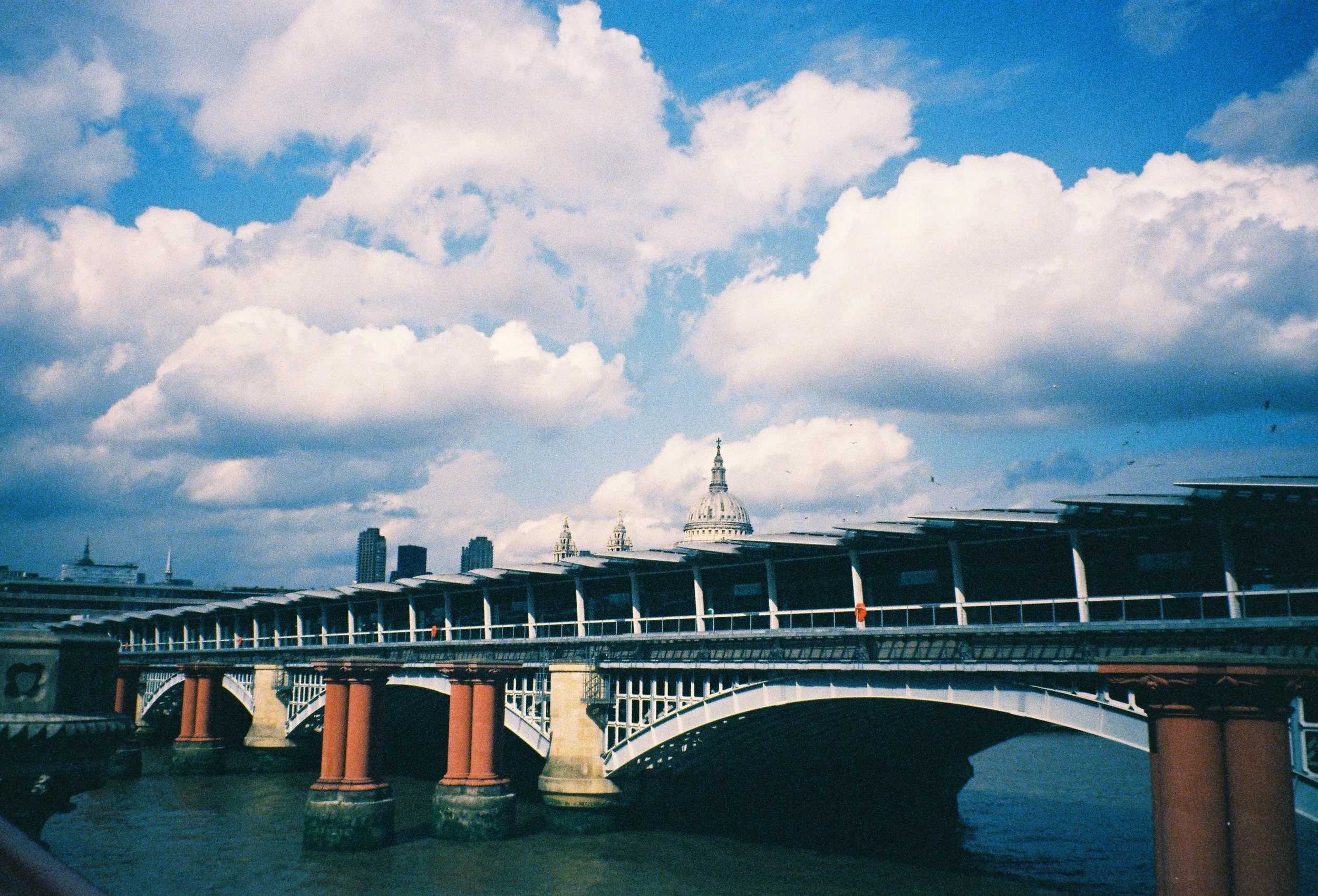 A cityscape with a bridge over a river, featuring classical architecture and a large domed building in the background under a partly cloudy sky.