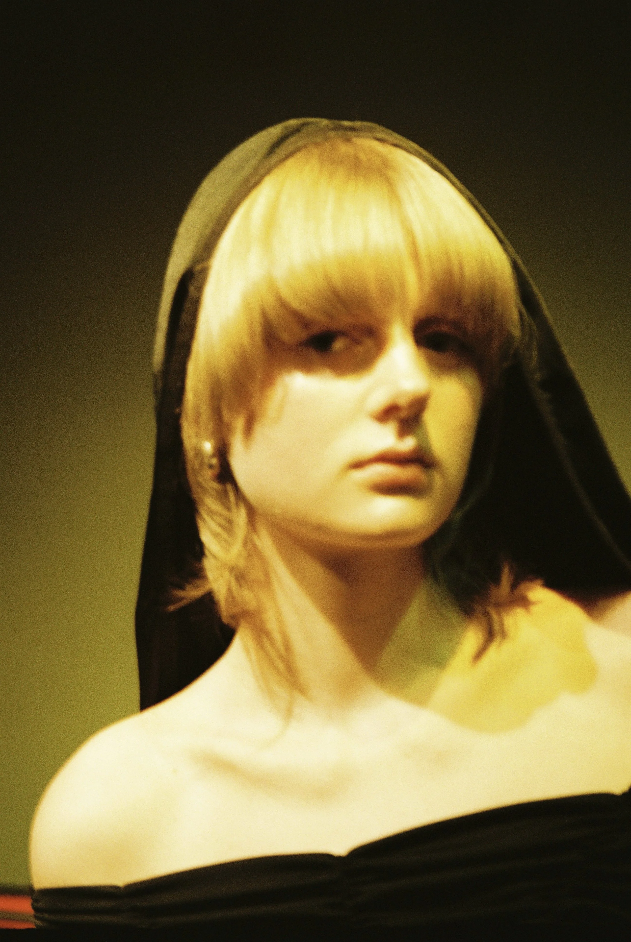 A close-up of a statue of a woman with blonde hair, wearing a black head covering and black clothing, with a neutral facial expression, set against a dark background.