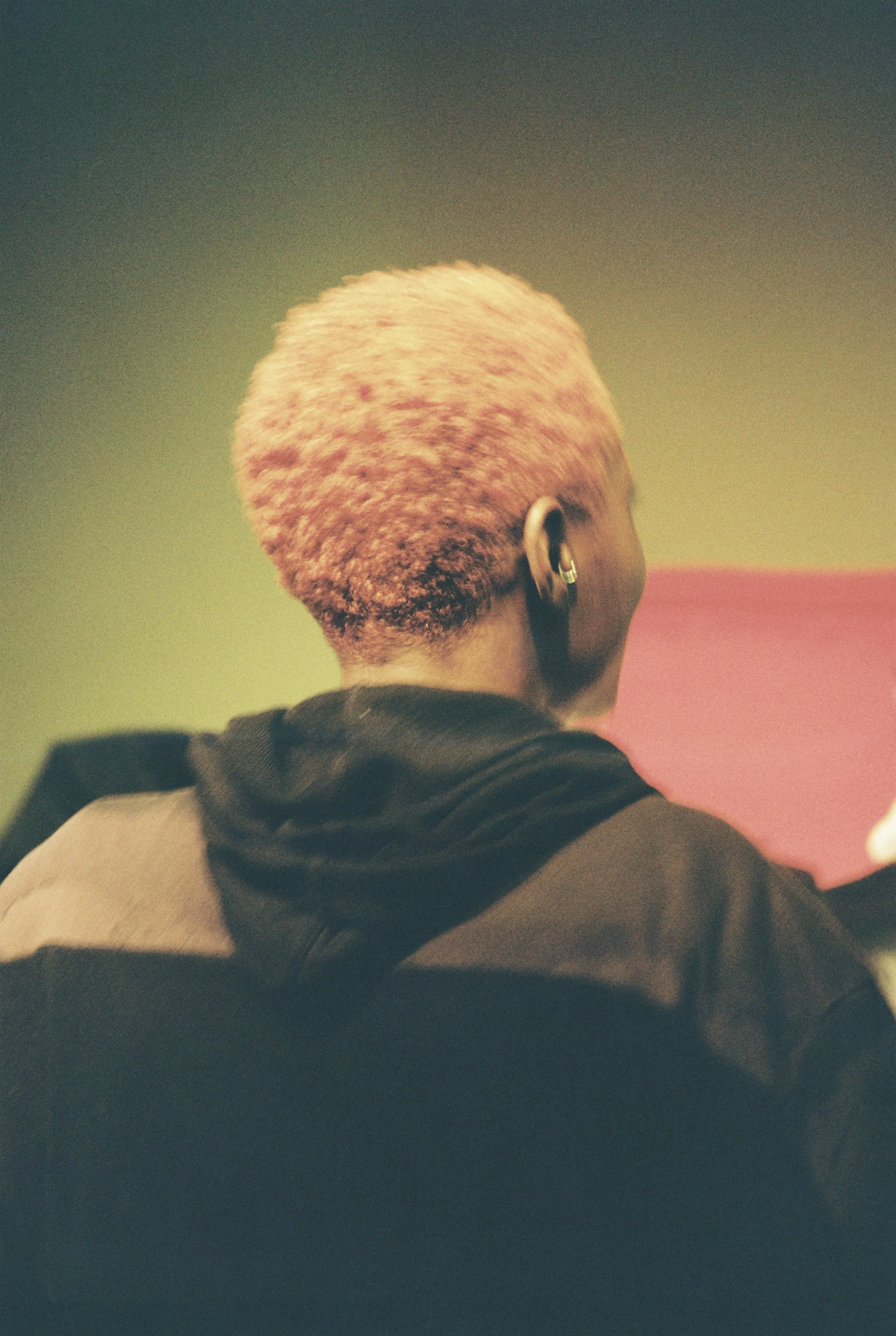 Back of a person with short, textured blonde hair, wearing a black jacket and large earrings, facing away from the camera in front of a pink and green background.