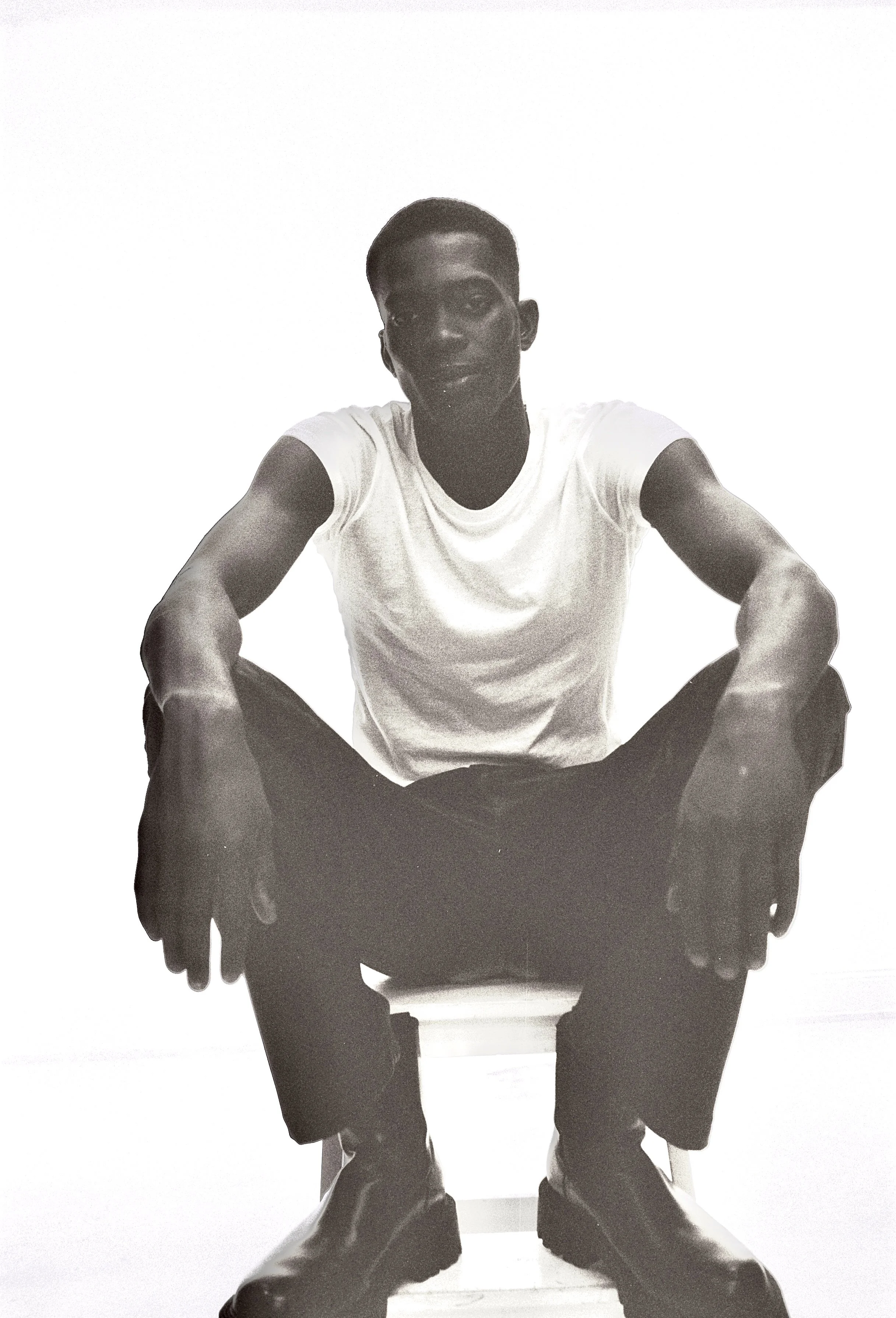 Black-and-white photo of a young man with short hair, sitting on a chair with legs apart, leaning forward slightly, hands resting on his knees, wearing a light-colored T-shirt and dark pants, against a plain background.