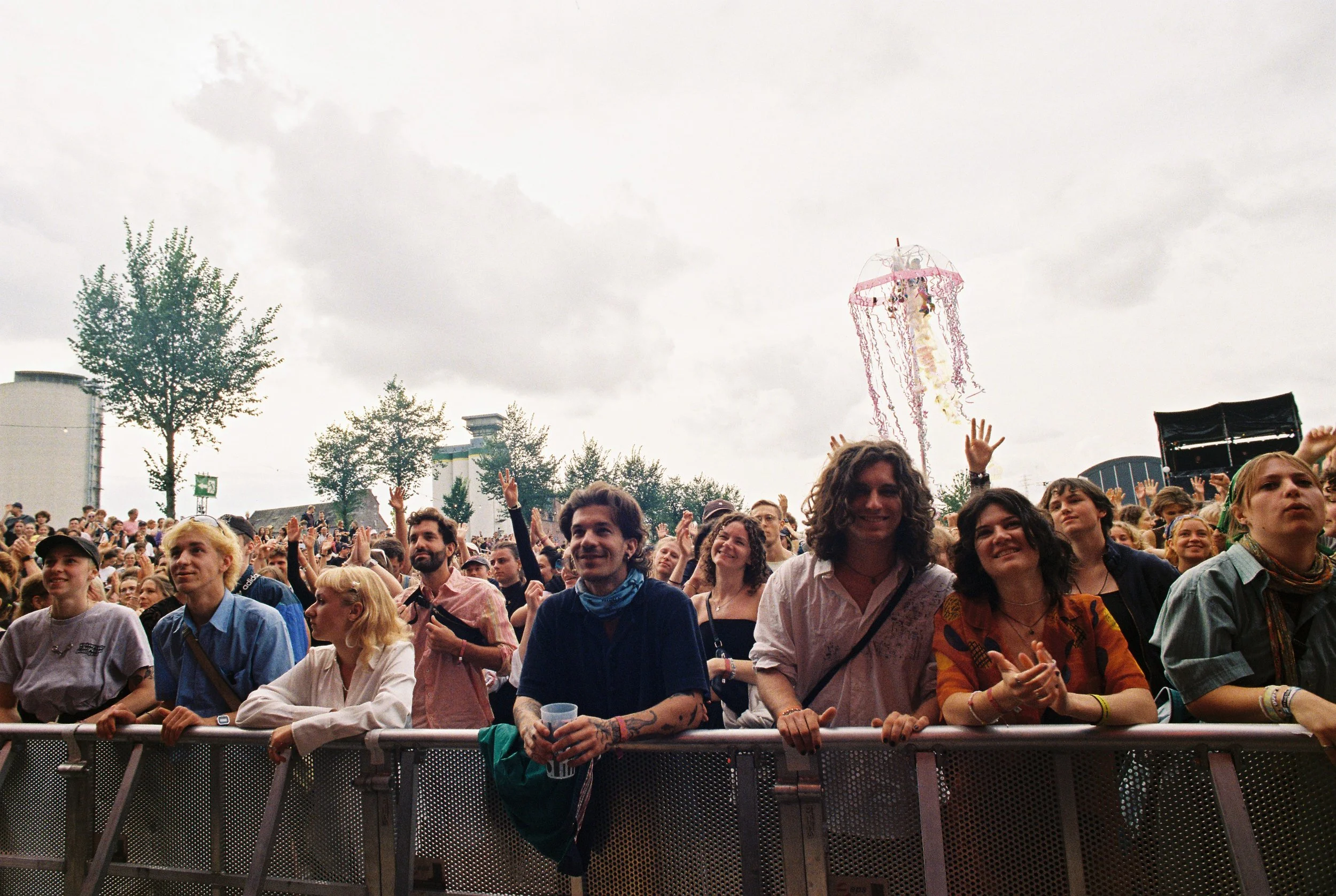 Crowd of people attending an outdoor concert or festival, standing behind a metal barricade, with some raising their hands and smiling. A clear umbrella with pink decorations is visible in the sky, and there are trees and buildings in the background 