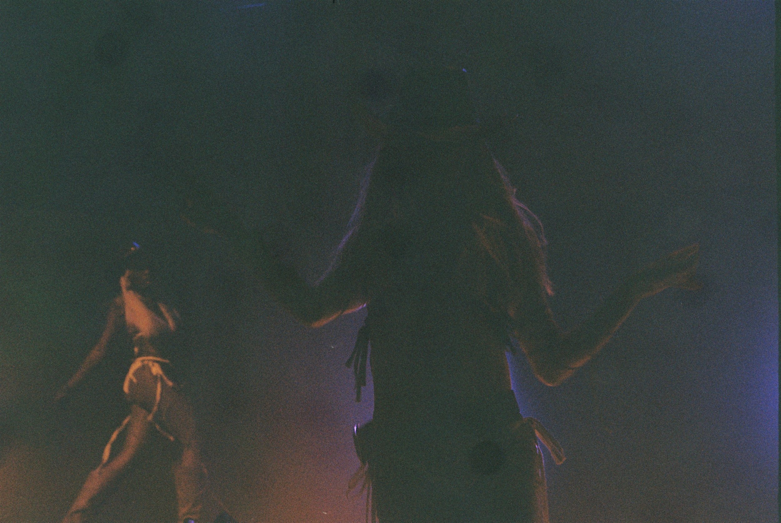 Silhouettes of people with raised hands in a dark, colorful environment, possibly at a concert or party, with purple and orange lighting.