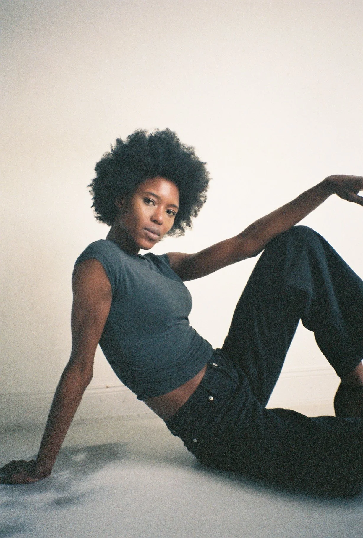 A woman with short curly hair sitting on the floor against a plain light-colored wall, wearing a dark gray t-shirt and black pants.
