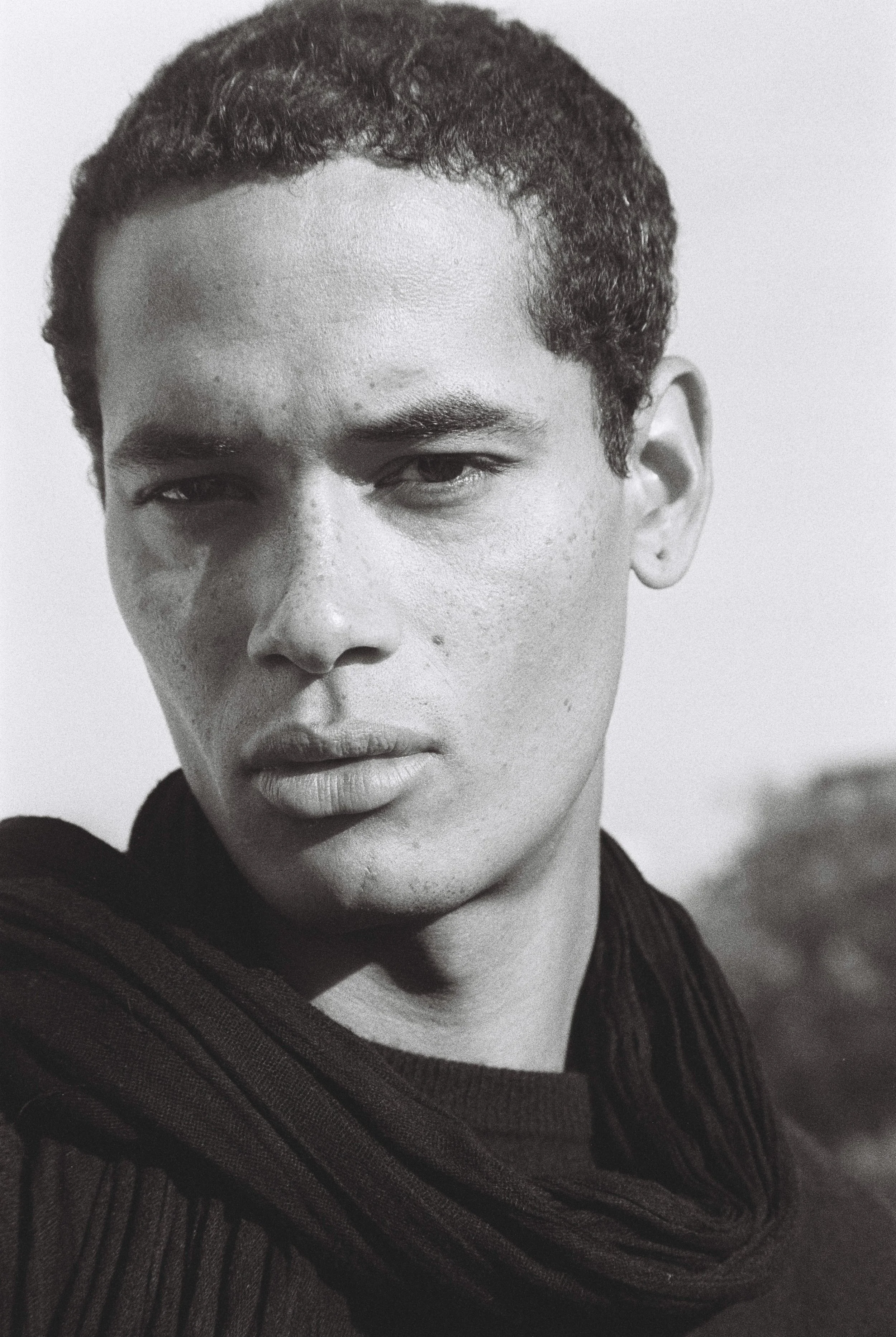 Close-up black and white photograph of a young man with short curly hair, wearing a dark textured sweater, looking slightly to the side.