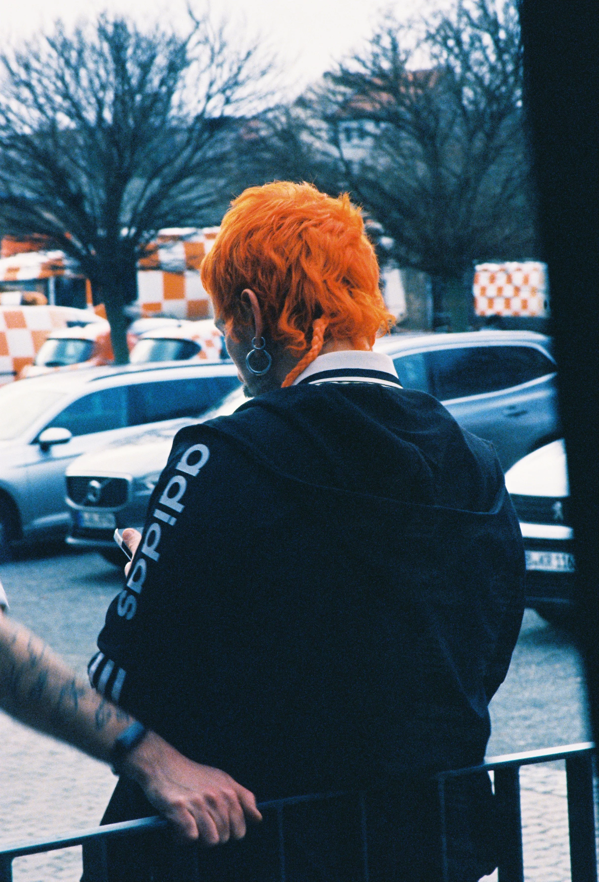 Person with bright orange hair, wearing earrings, dressed in a black jacket, standing by a metal railing near cars and leafless trees, holding a phone.