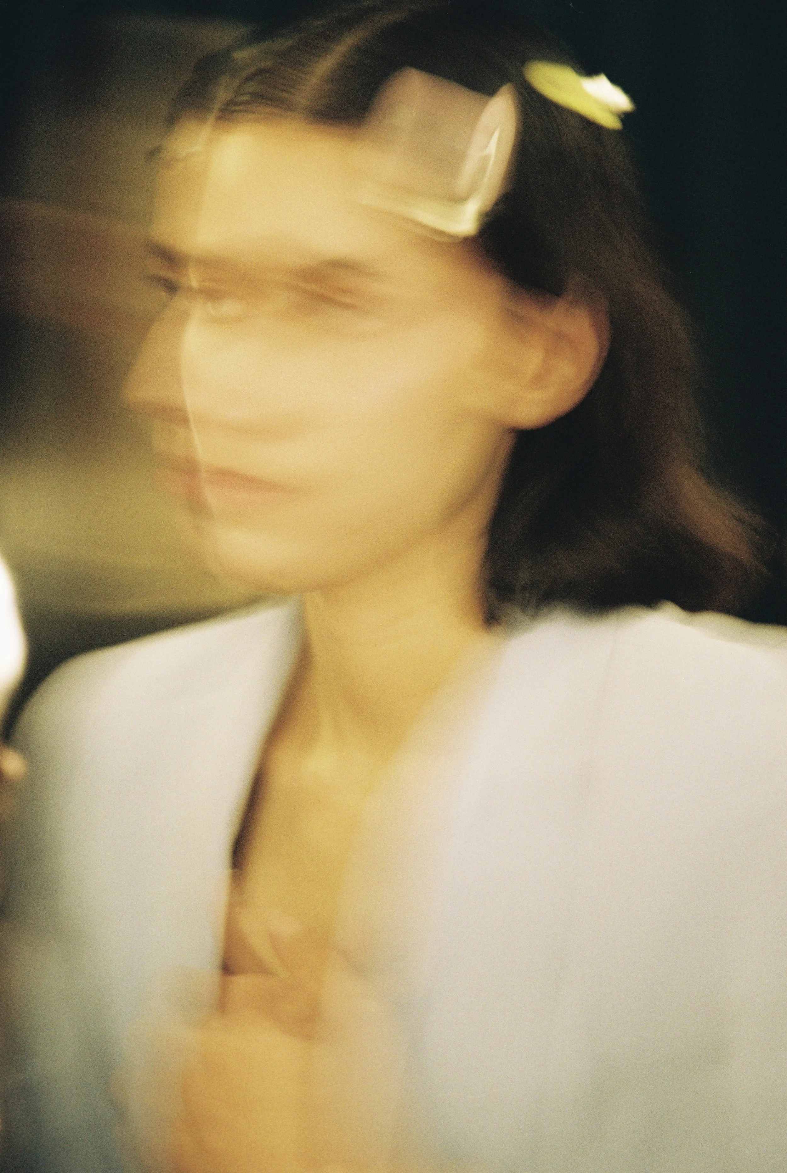 A woman with dark brown hair tied back, wearing large sunglasses resting on her head and a white top, gazing downward in a soft-focused portrait.