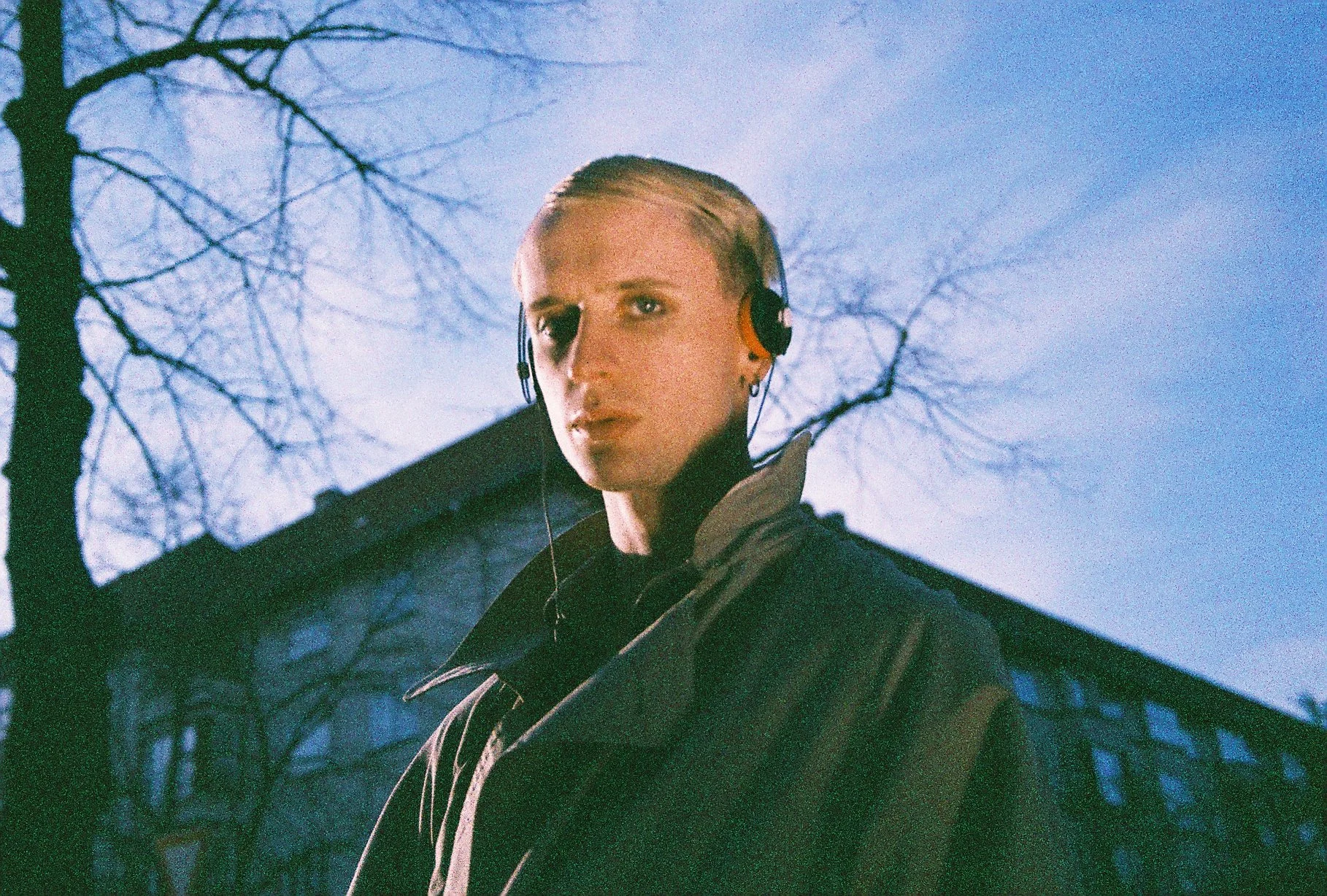 A young person with short blond hair, wearing headphones and a dark jacket, looking at the camera against a background of leafless trees and a blue sky.