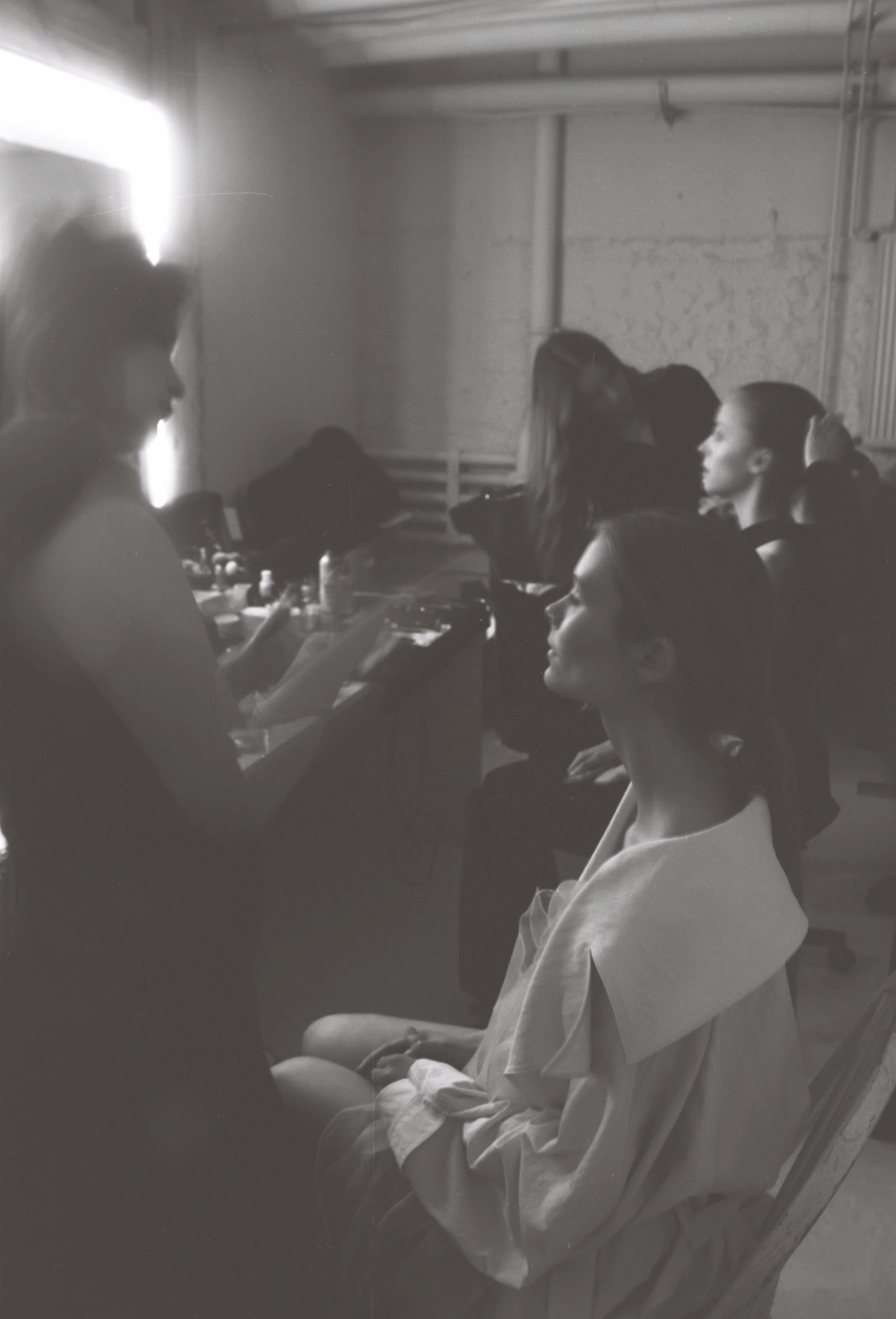 A black and white photo of a young person with short hair sitting on a hospital or clinic bed, wearing a hospital gown, with some medical equipment or supplies beside them. Other people are present in the room, possibly medical staff or visitors, wit