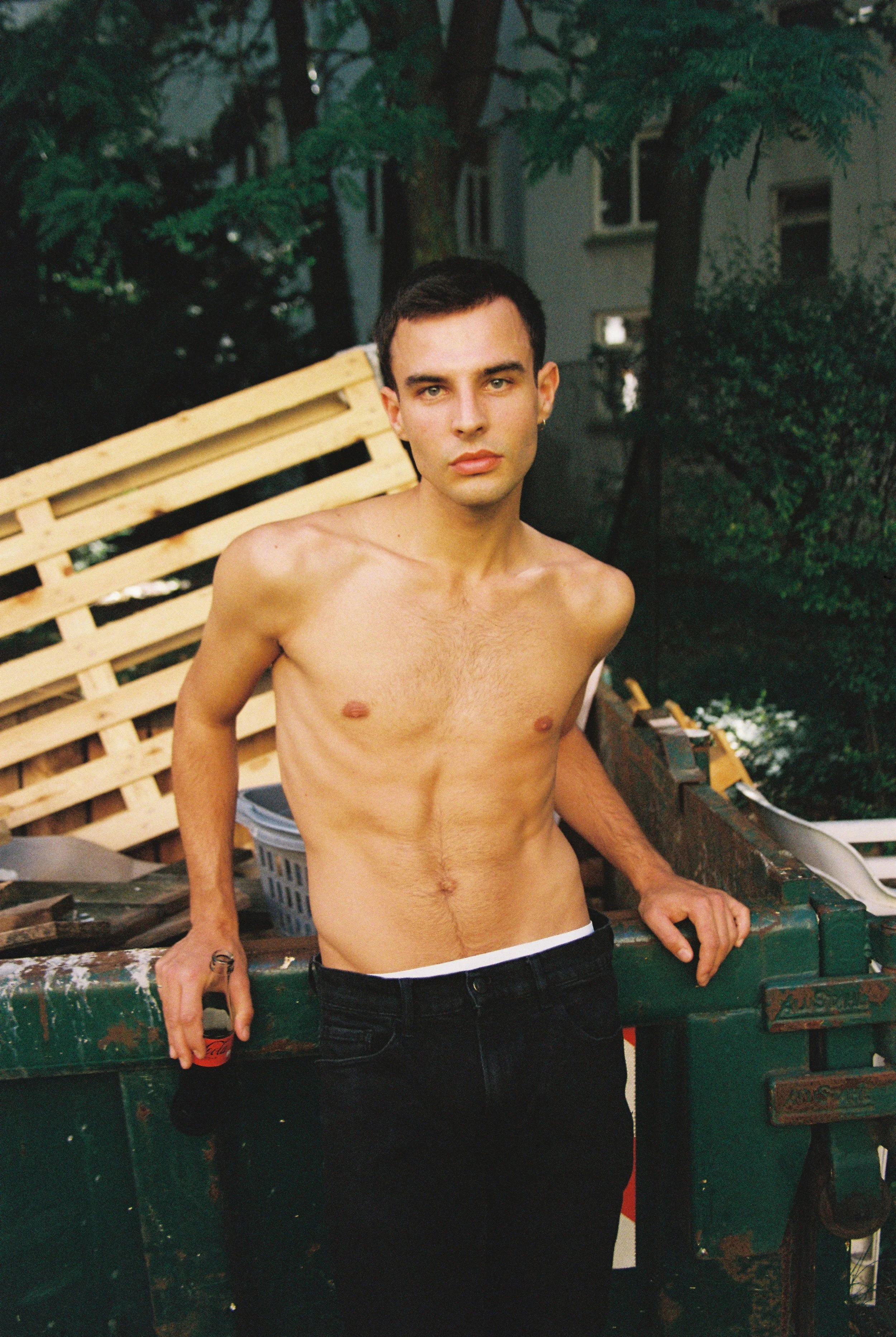 A shirtless young man with dark hair and light skin posing outdoors in a backyard setting, leaning on a large green metal trash bin.