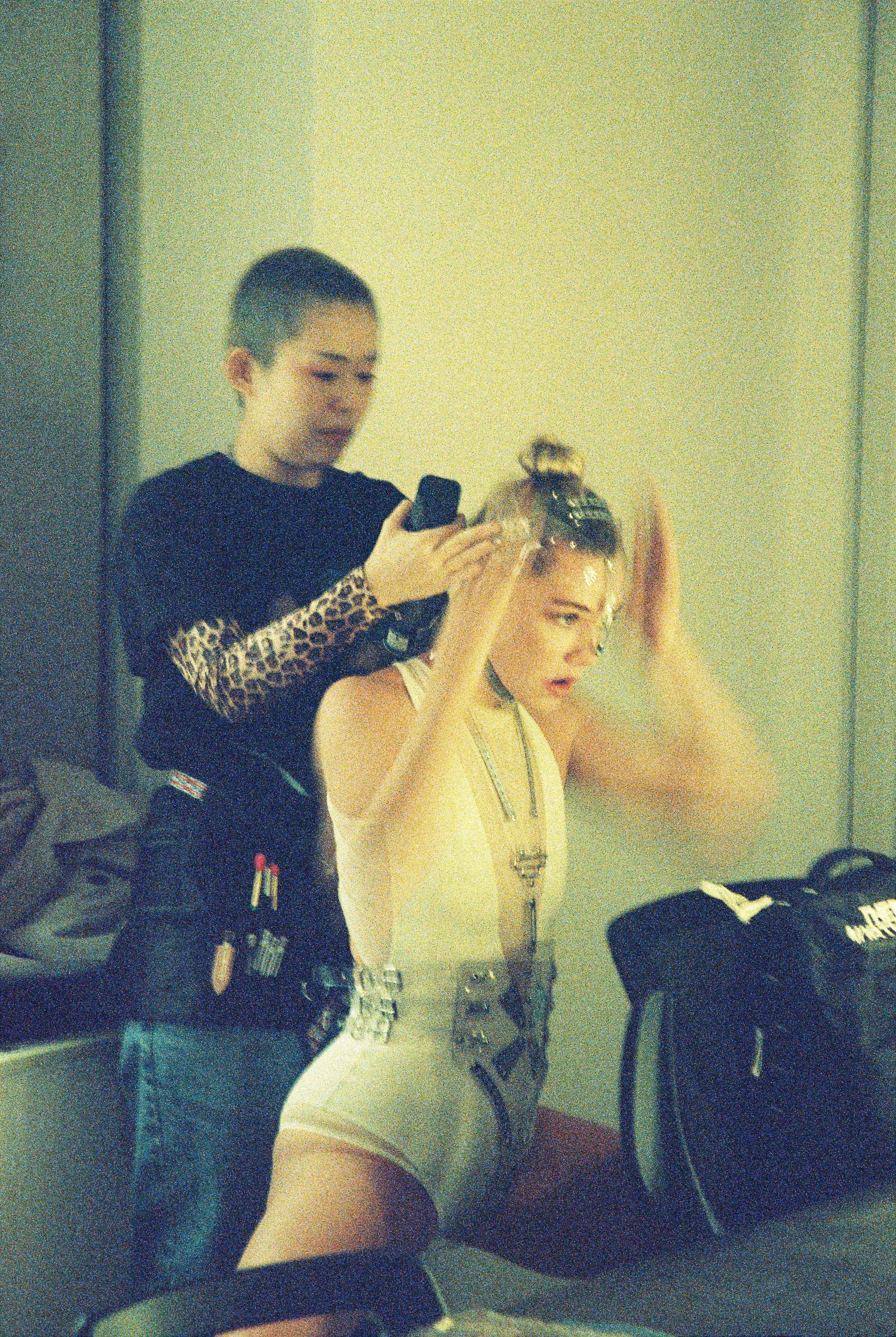 A young woman with short blonde hair dressed in a revealing white outfit, is getting her hair styled by another woman with short dark hair in a black top. The setting appears to be backstage or a dressing room, with makeup and styling supplies around
