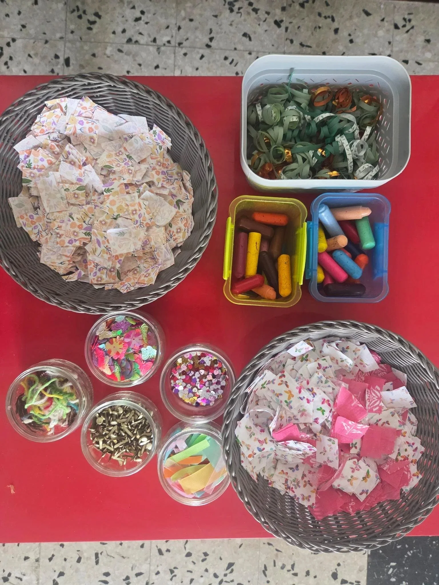 Papier décoratif, confettis, rubans et des petits objets colorés, disposés dans plusieurs petits bols et paniers sur une table rouge.