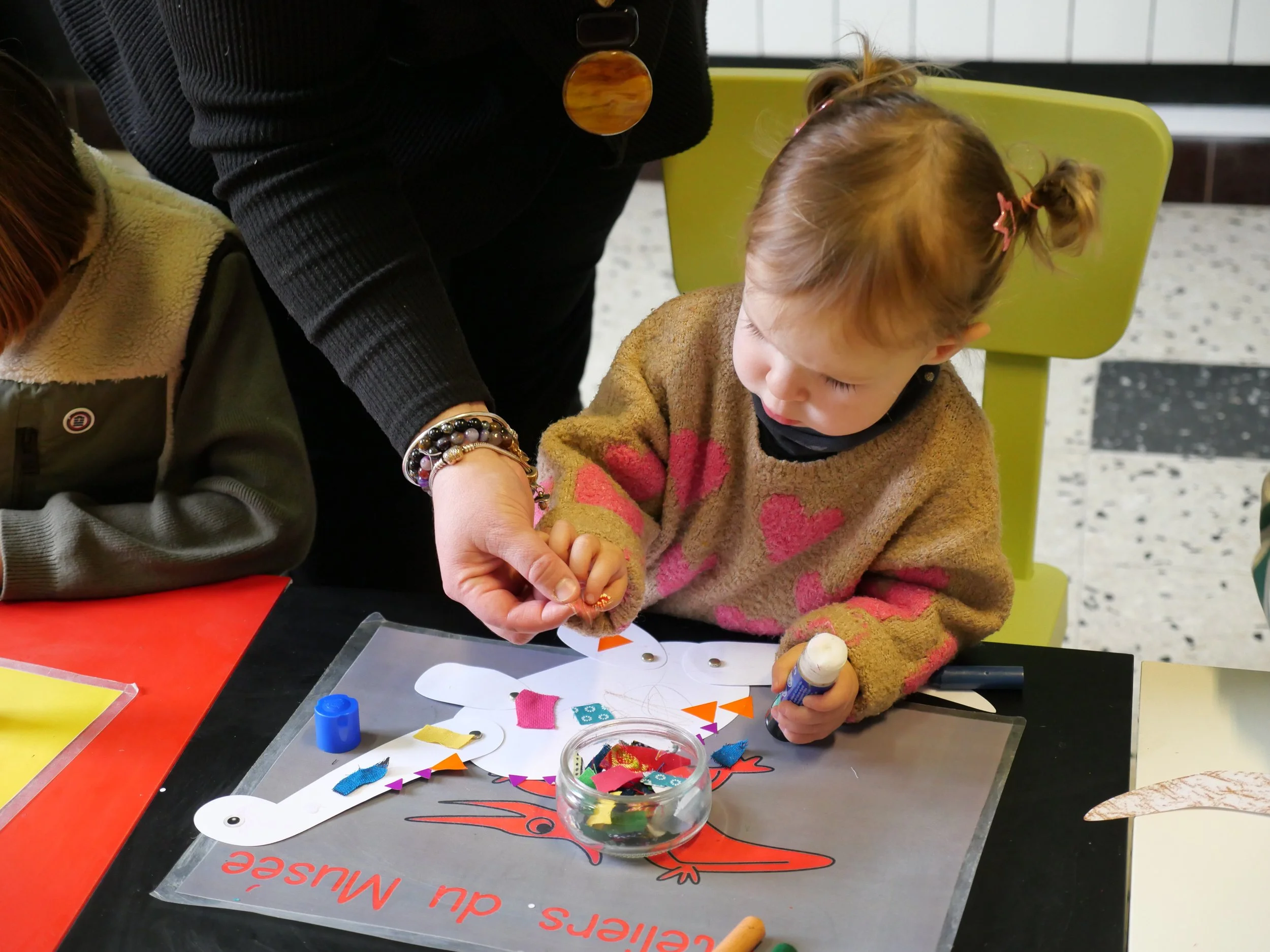 Une petite fille fait une activité créative avec des matériaux colorés sur une table. On voit du papier de différentes couleurs, des colle, des petits morceaux de tissu ou de papier, possiblement pour une création artistique ou un projet scolaire.