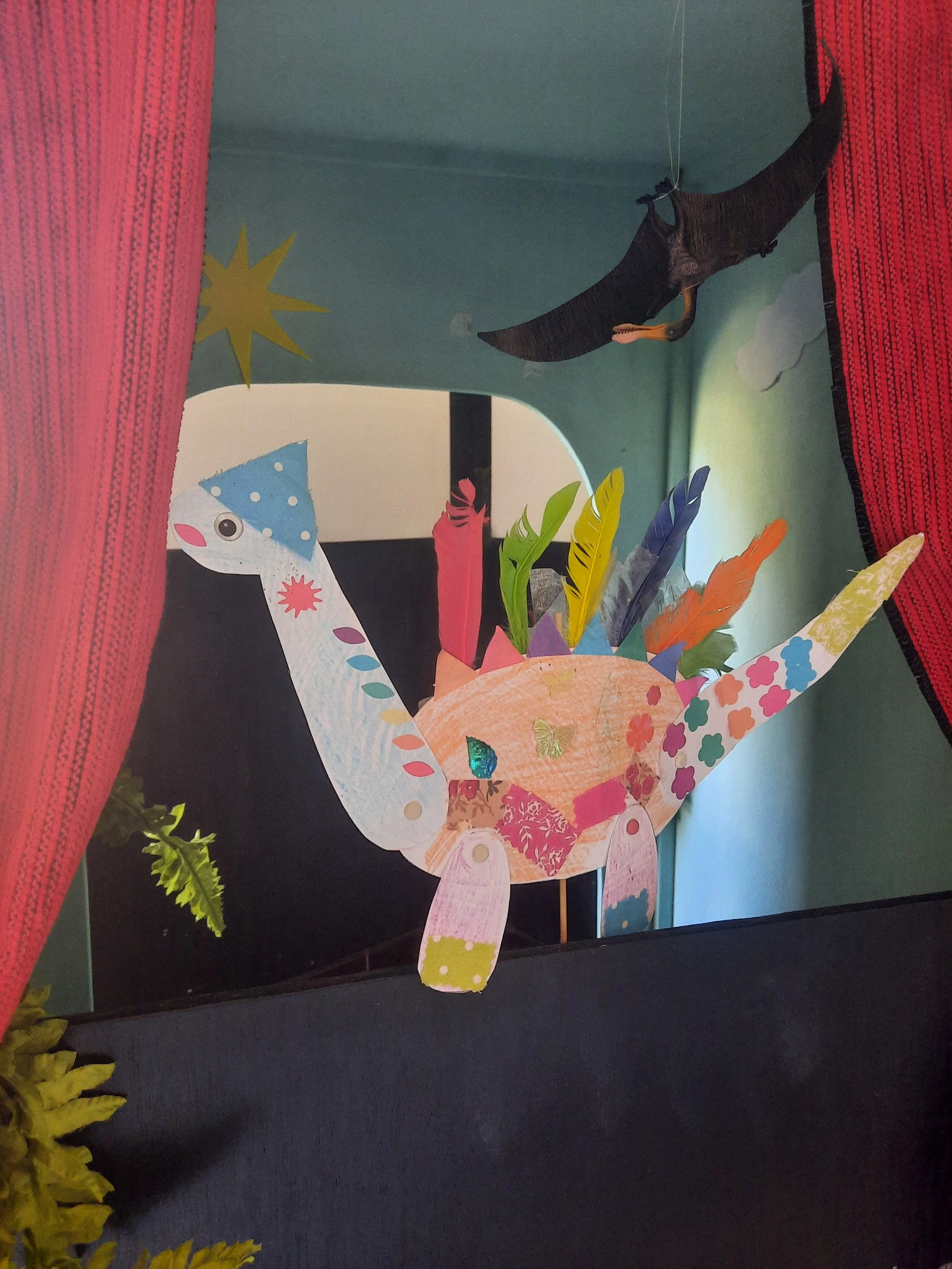 Marionnette d'un dinosaure coloré avec des plumes en papier multicolores, entouré de décorations de fête et de rideaux rouges.