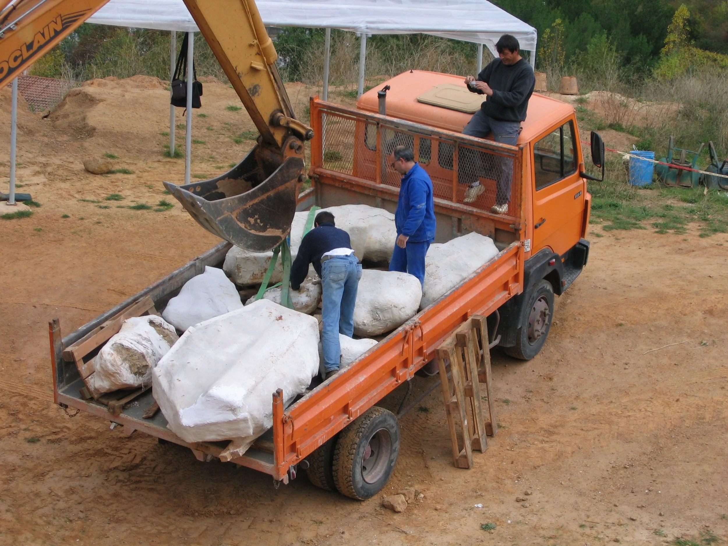 Un camion orange transportant des blocs de pierre. Trois hommes travaillent autour du camion, un d'eux est debout à côté, un autre est à l'intérieur chargeant les blocs, et un troisième est assis sur la cabine du camion. Il y a un terrain en terre avec quelques herbes et un espace extérieur arboré en arrière-plan.