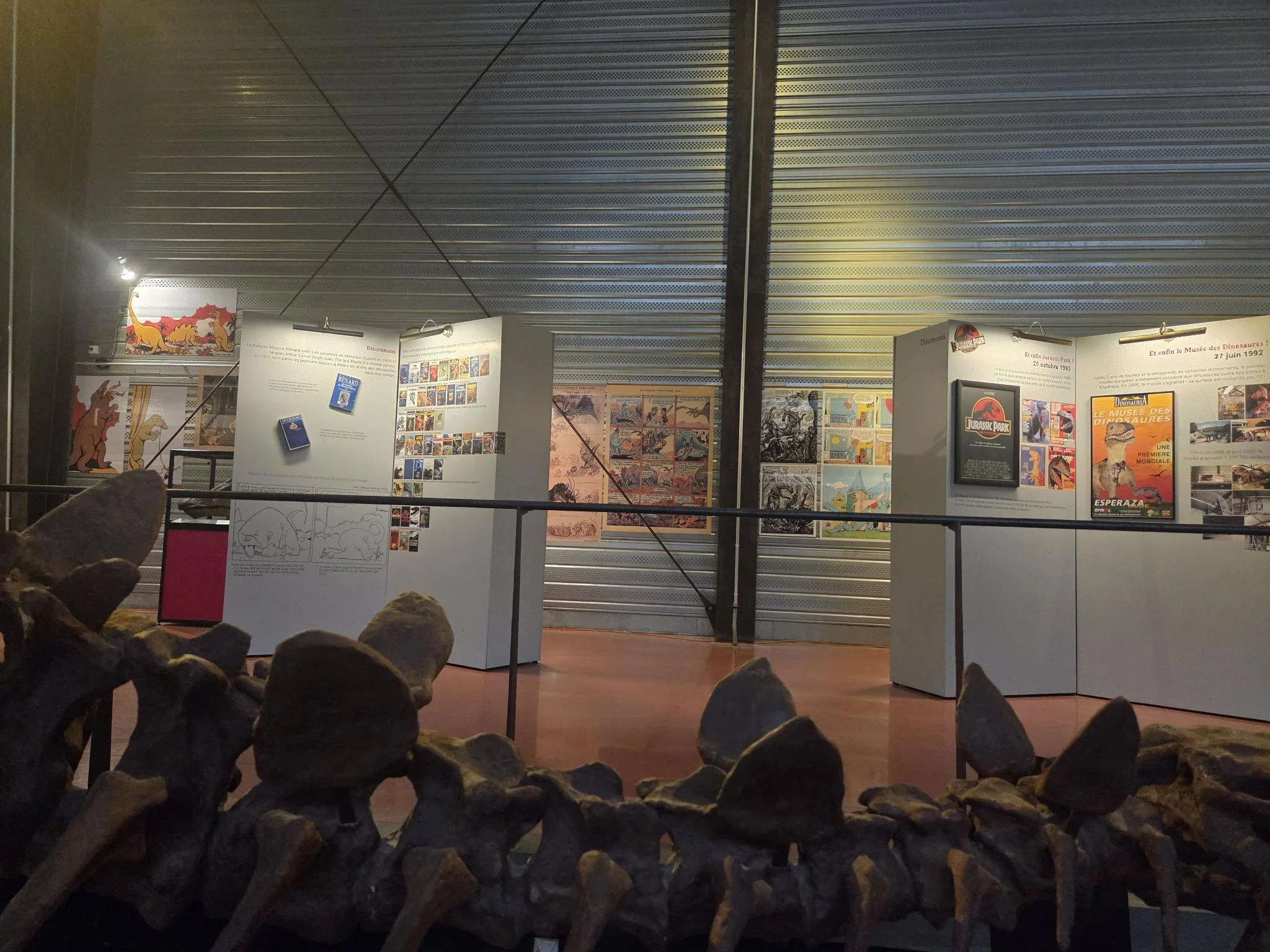 Une exposition sur les dinosaures avec des panneaux informatifs et affiches. Présence d'une maquette ou panneau illustrant un dinosaure, et des informations historiques. La scène semble être dans un musée ou une lieu d'exposition.