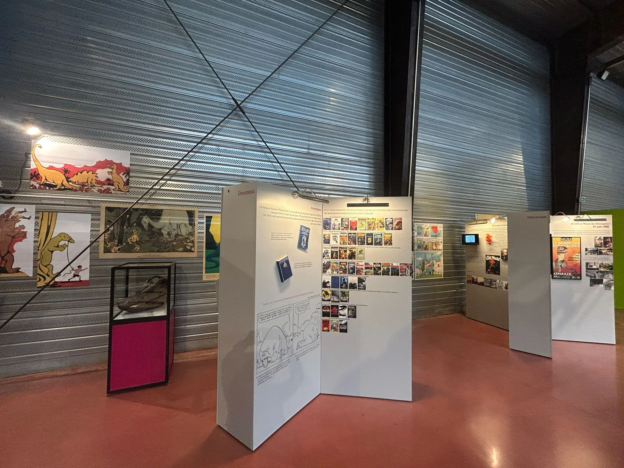 Une exposition intérieure avec plusieurs panneaux d'informations, affiches, et illustrations sur des dinosaures, dans une salle avec mur en métal et éclairage modéré.
