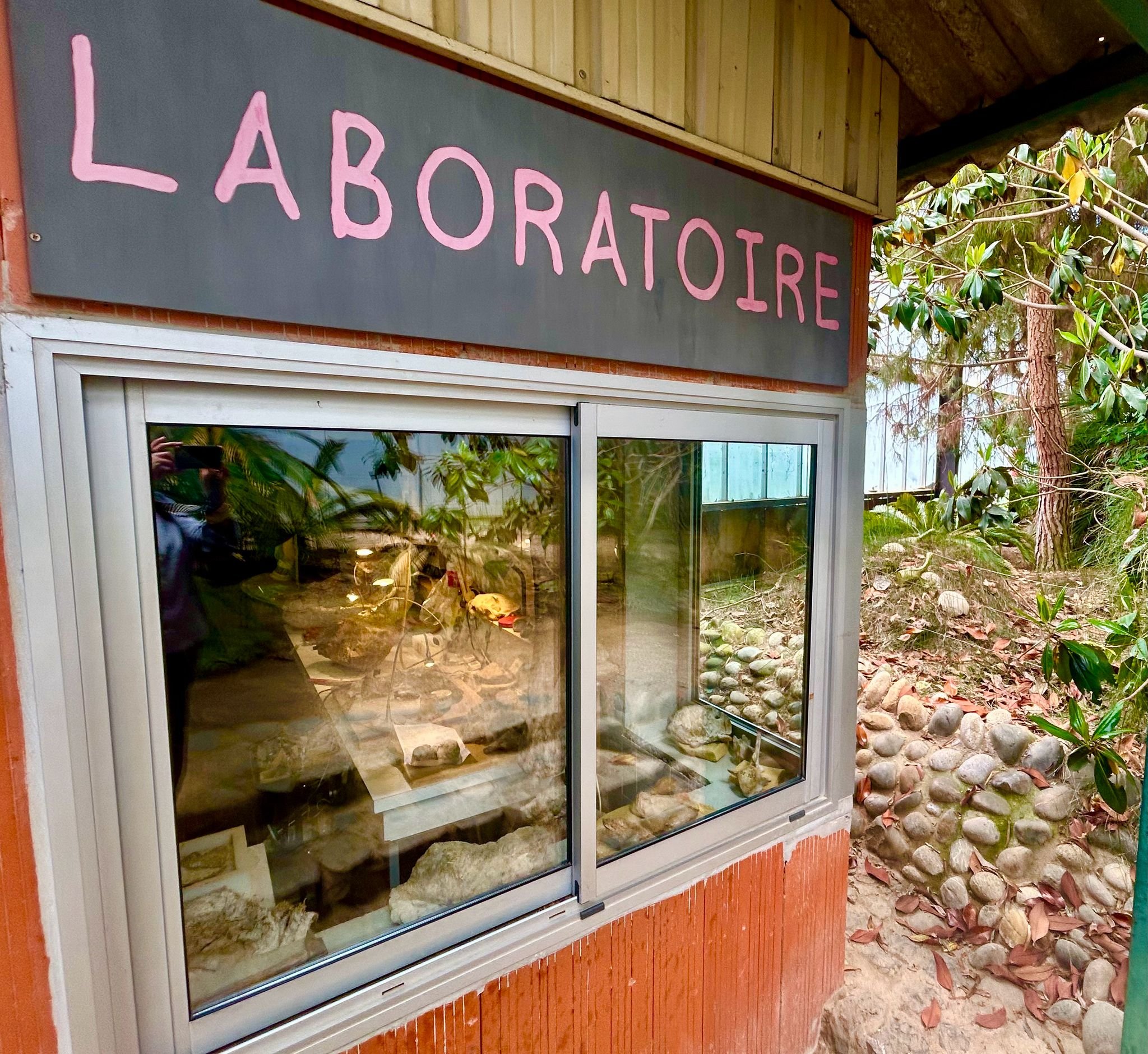 Une devanture de boutique ou de stand avec un panneau indiquant 'LABORATORIE' écrit en lettres roses sur fond gris, entourée de végétation et de pierres.