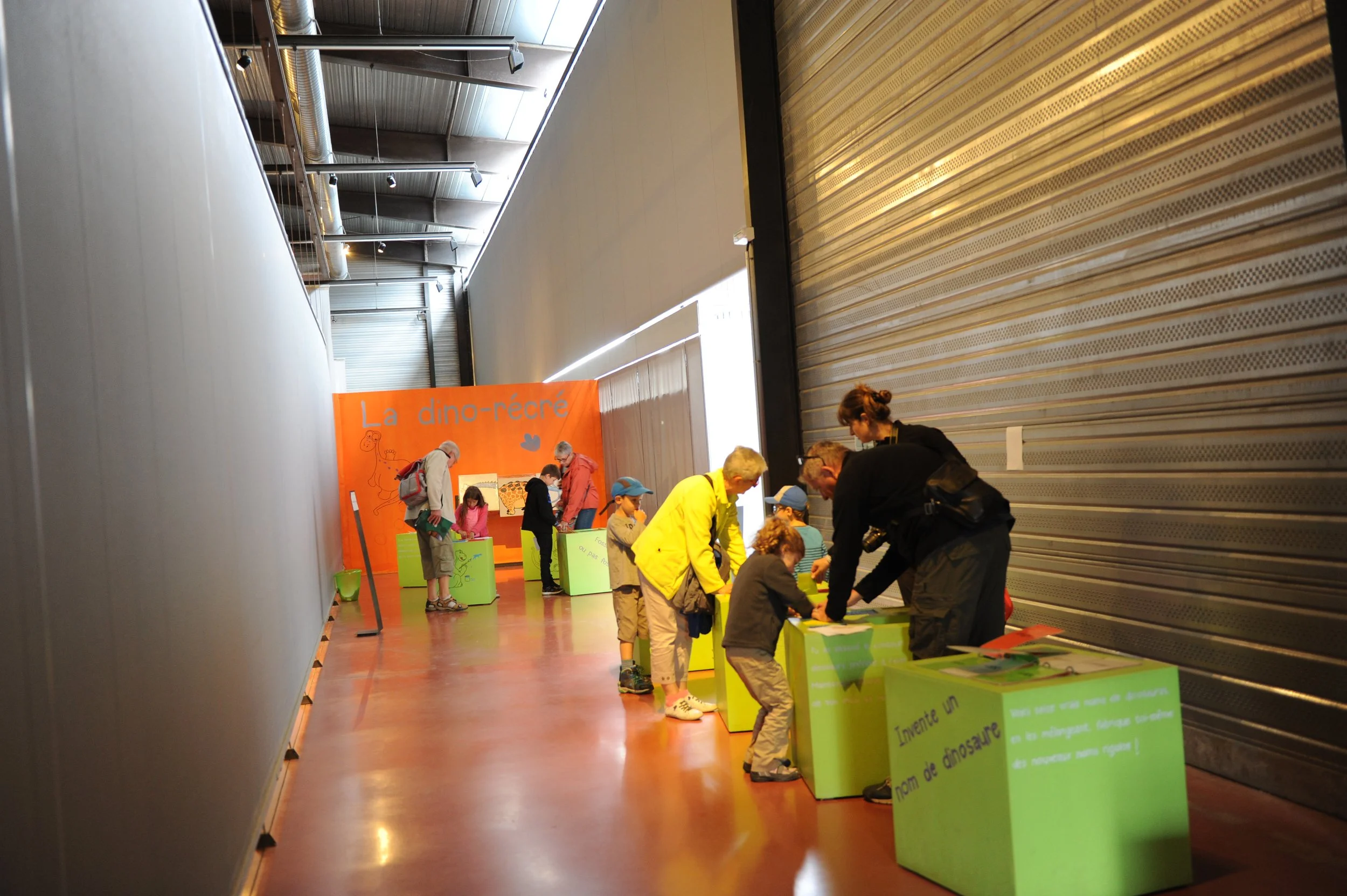 Une activité éducative pour enfants avec des tables colorées dans un centre d'exposition, ayant un décor orange avec le texte 'La dino-recré' et sans personnes visibles.