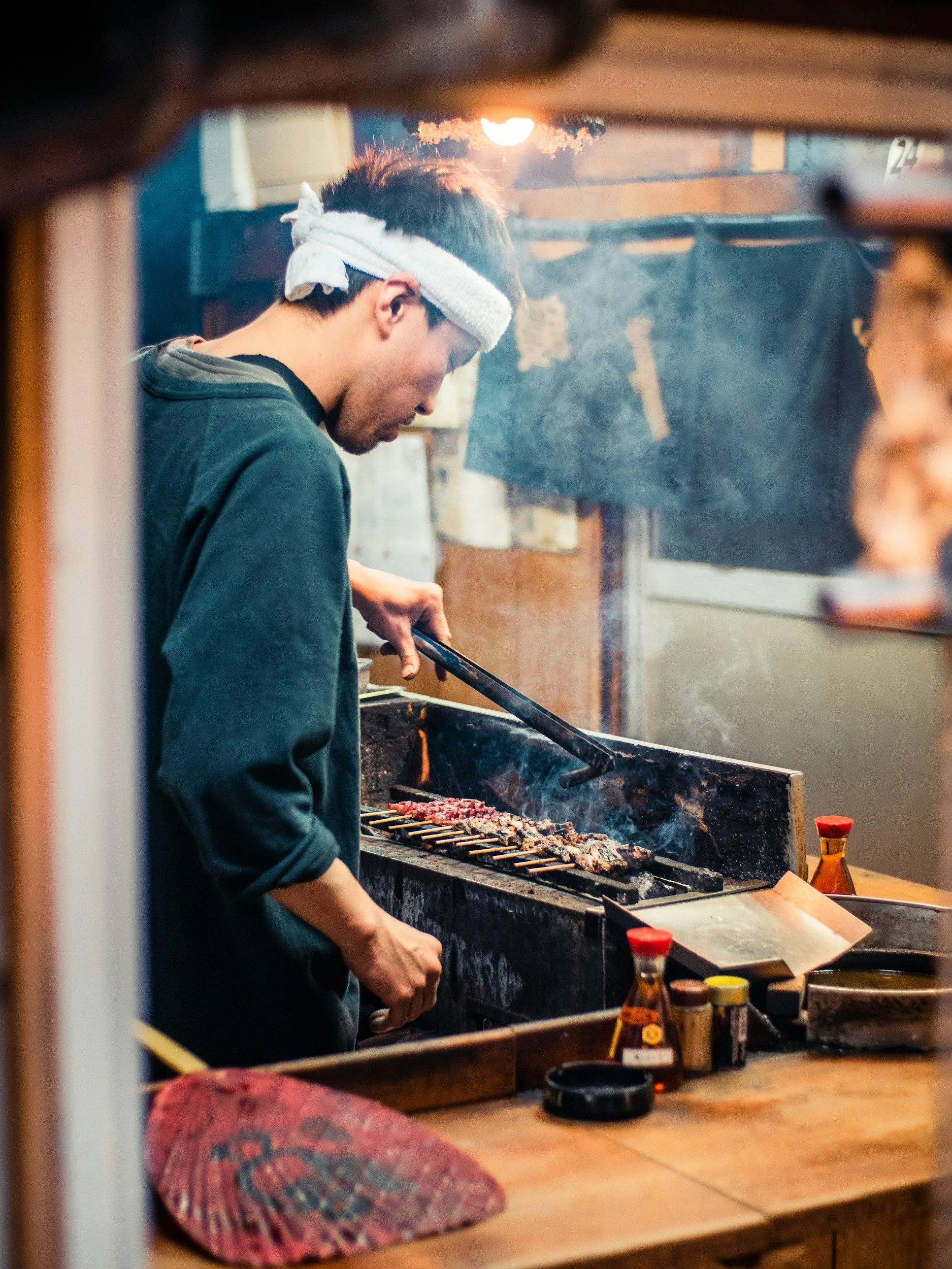 A Japanese cook wearing a white towel wrapped around their head is grilling multiple skewers of meat on a barbecue grill in a robata restaurant setting.