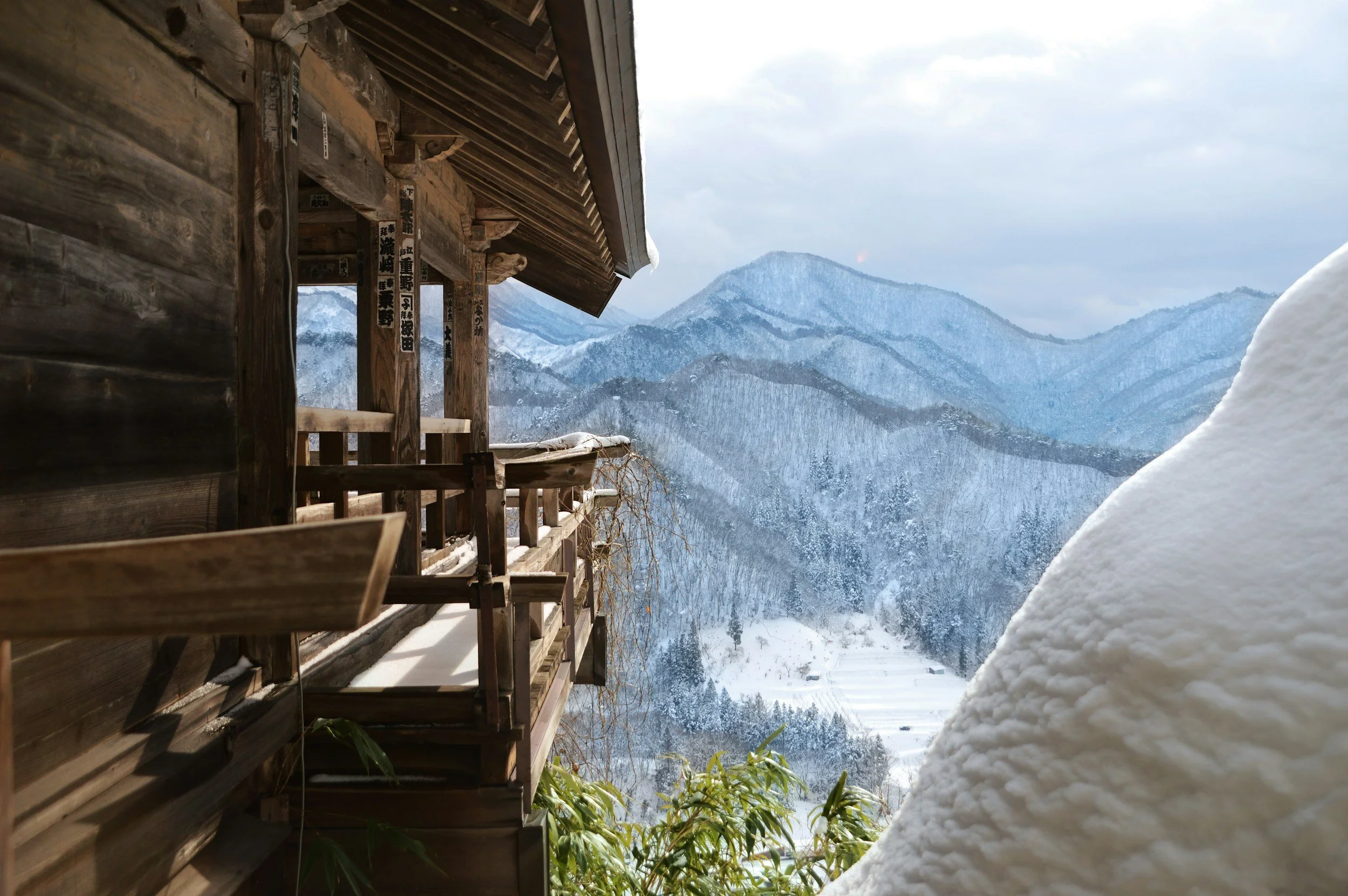 Wooden balcony with snow, overlooking snow-covered mountainous landscape under cloudy sky.