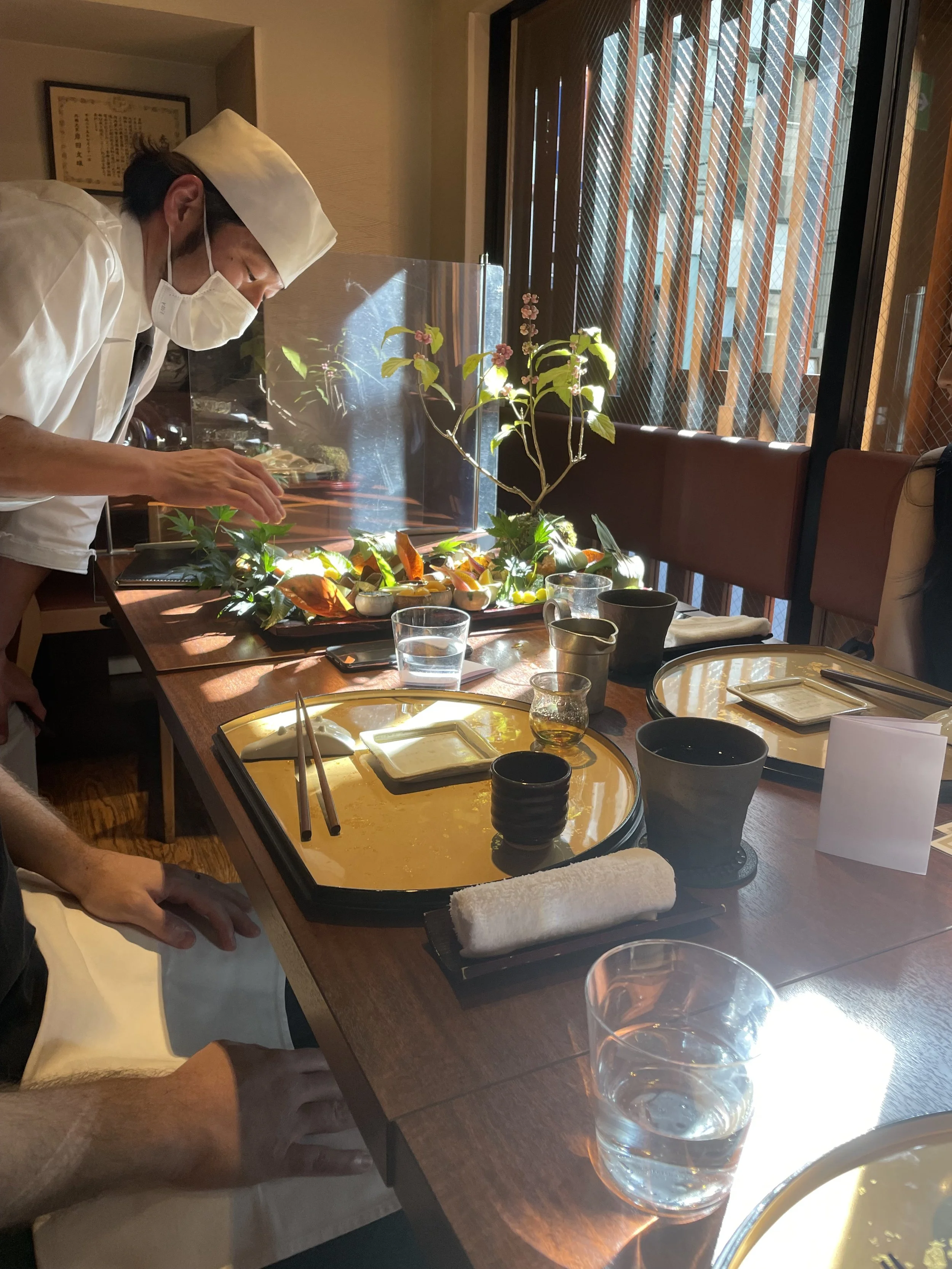 A Japanese chef wearing a white uniform, and hat is setting a table in a Izakaya restaurant. The table has a small tray with traditional Japanese food dishes.