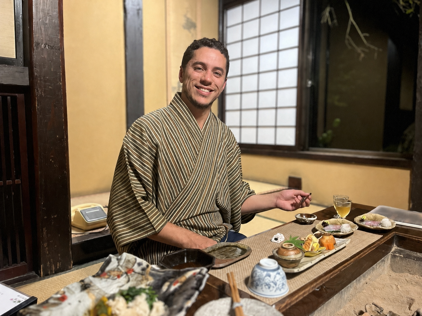 A smiling man in traditional Japanese attire sitting at a low dining table with various Japanese dishes, tea, and tableware in a Japanese-style room.