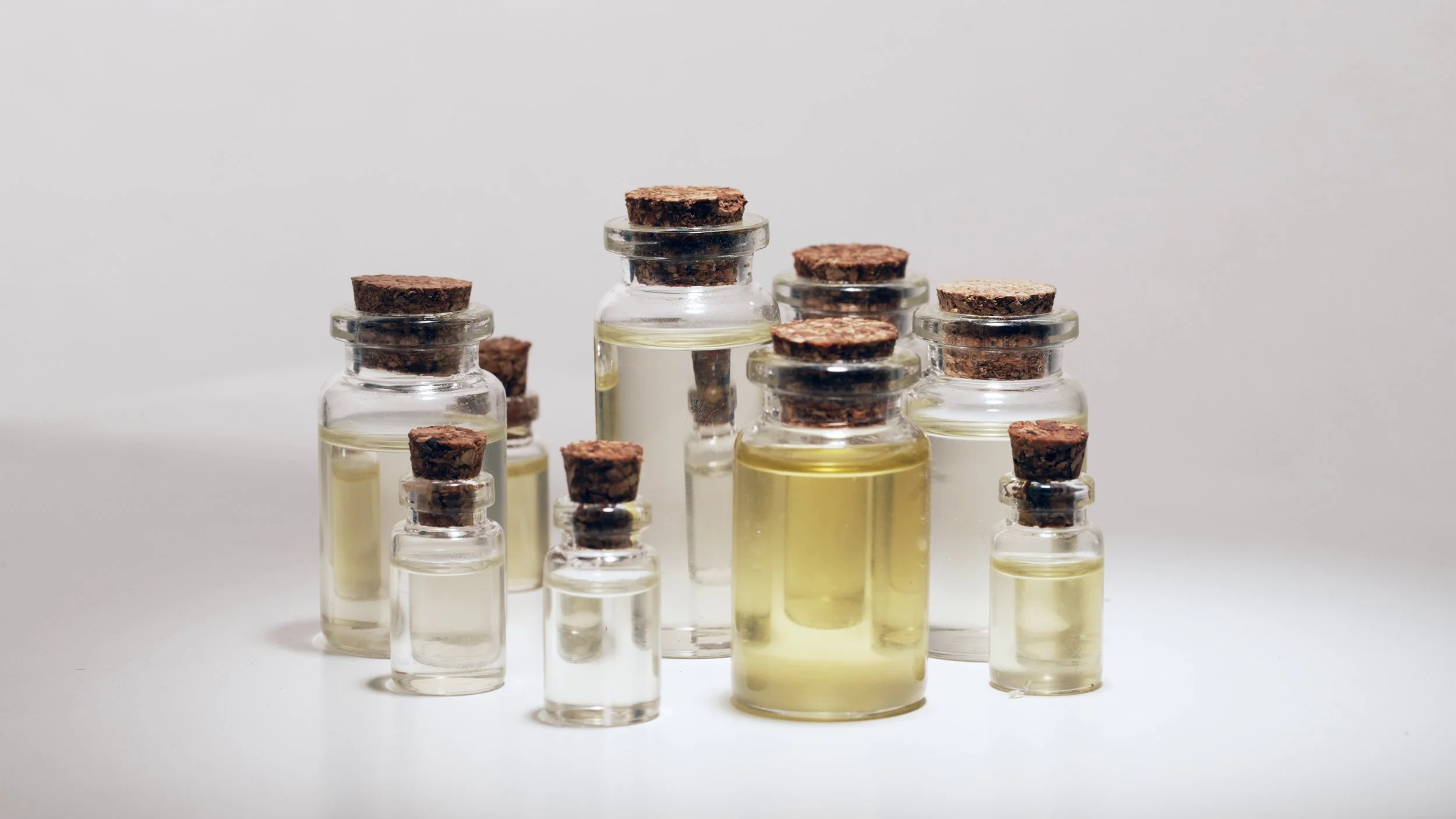 Multiple small glass bottles with cork stoppers containing a clear yellow liquid, arranged on a white surface against a plain background.