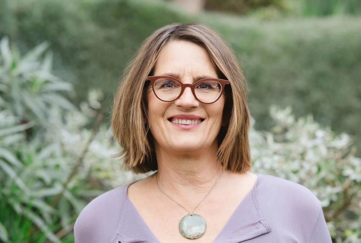A smiling middle-aged woman with shoulder-length light brown hair, wearing glasses and a purple top, outdoors with greenery in the background.