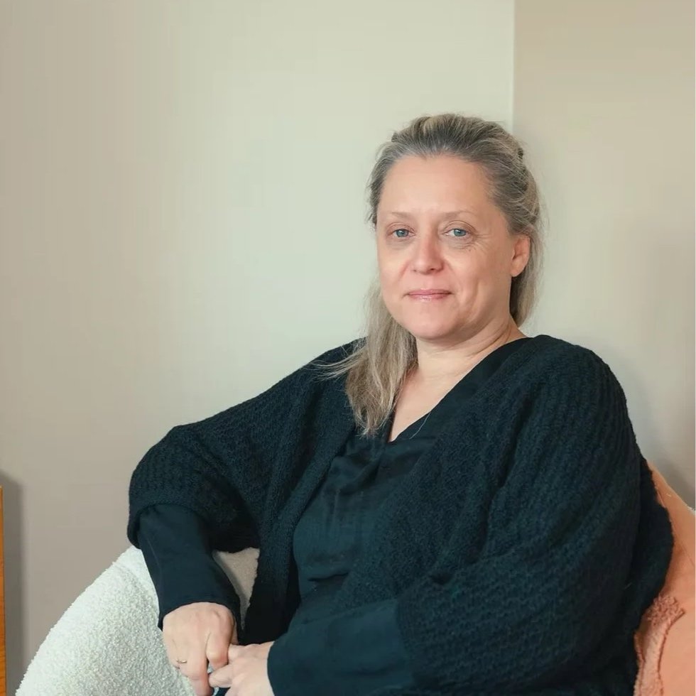 Een vrouw met grijs haar die in een zwarte jas zit, zittend op een bank in een kamer met een witte muur.