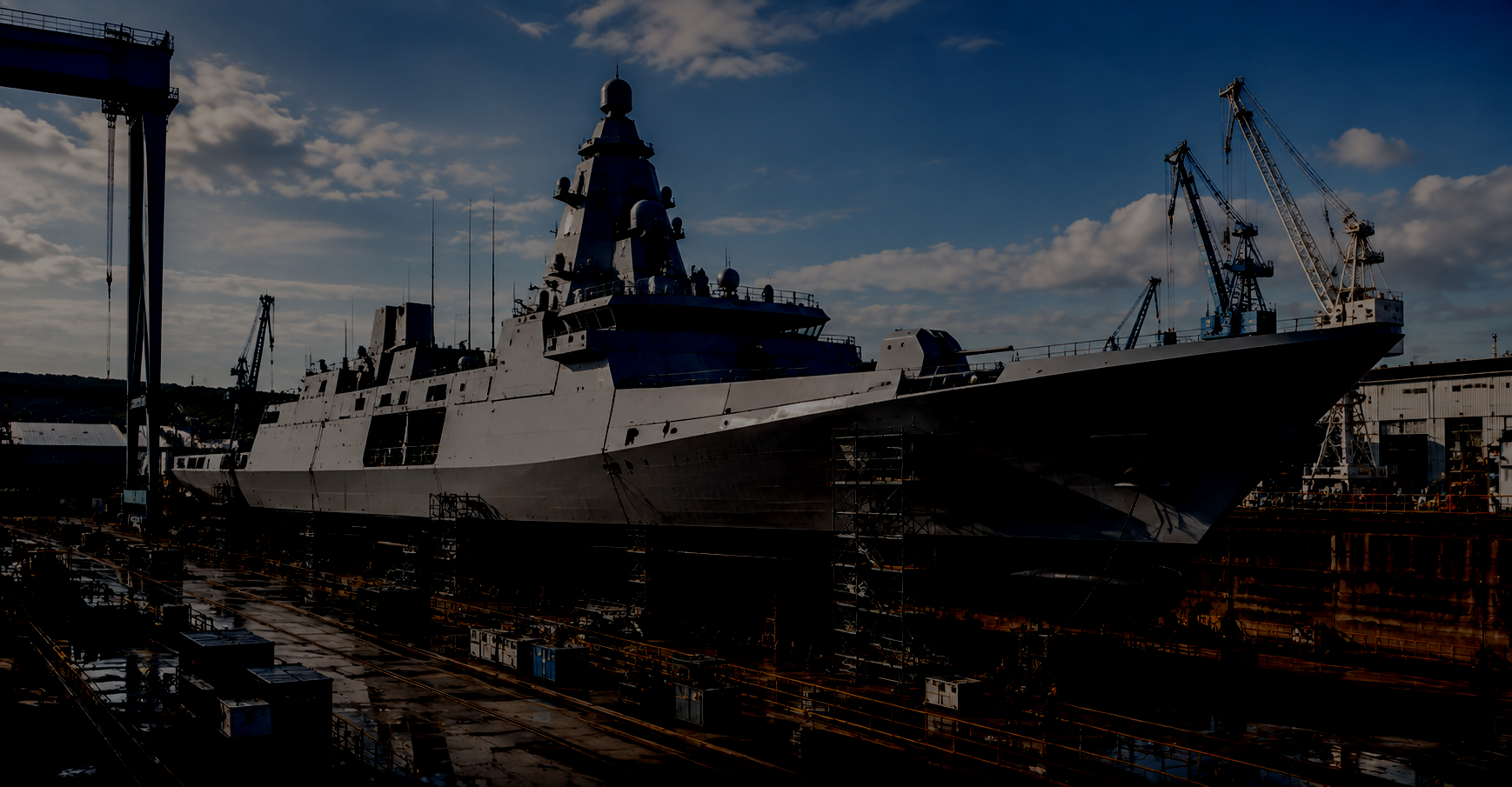 Ein großes Kriegsschiff liegt im Trockendock, umgeben von Kräne und Baugerüste, bei Sonnenuntergang oder -aufgang.