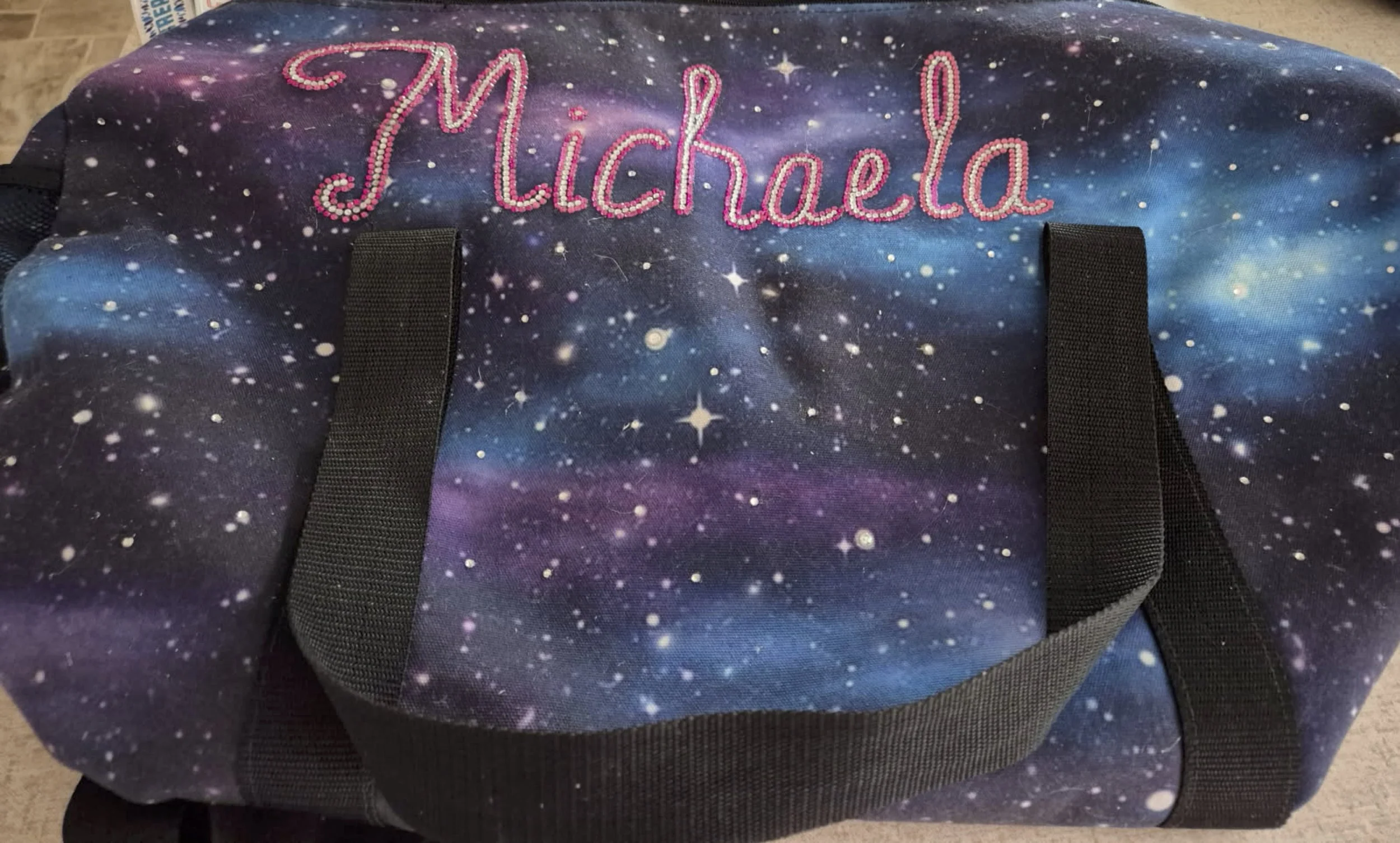 Bag with a galaxy print design featuring stars and cosmic patterns, with the name 'Michaela' in pink and silver diamonds on the top.