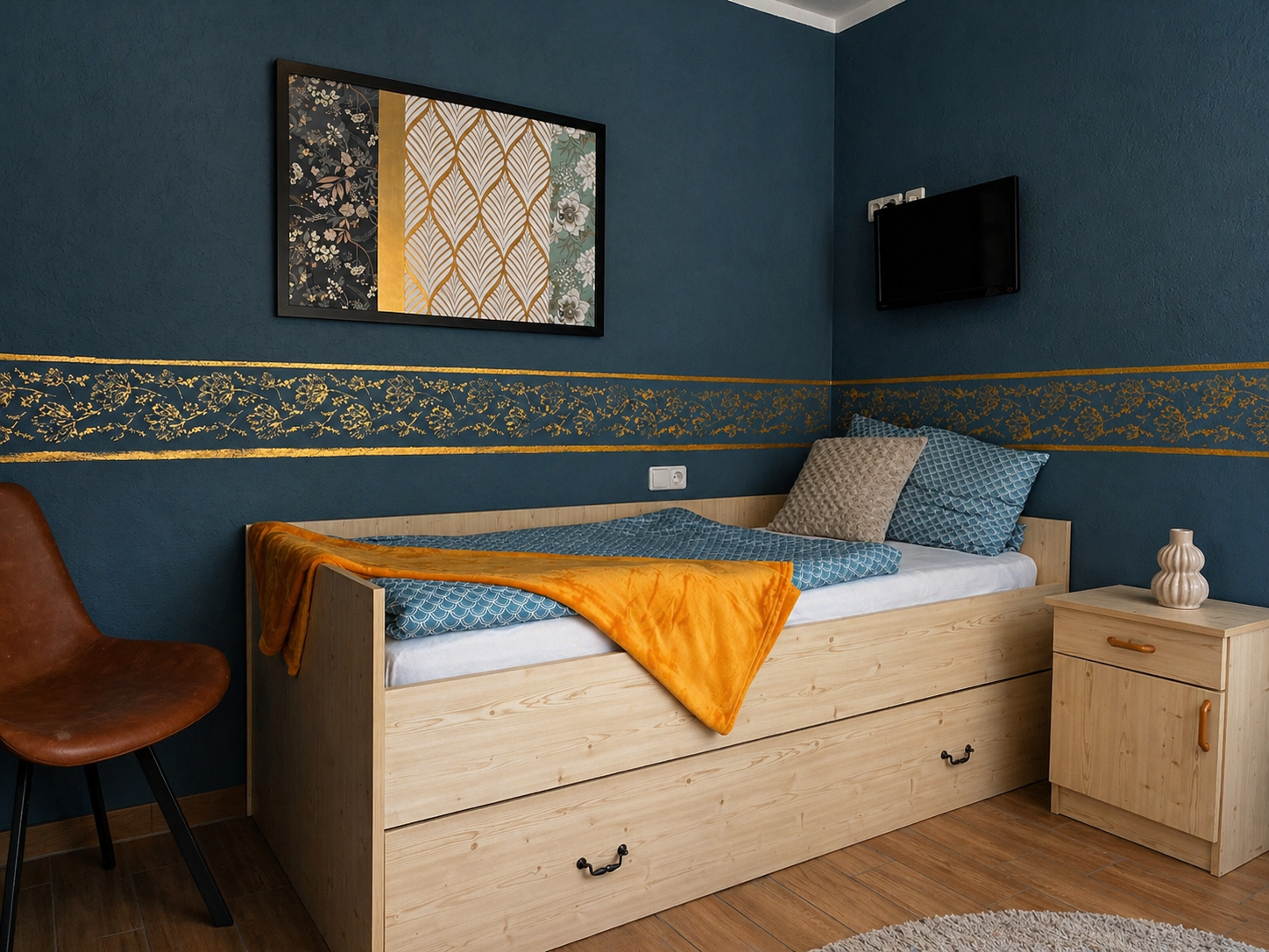 Ein Schlafzimmer mit blauer Wandfarbe, einem Holzbett mit blauen Kissen, einer orangefarbenen Decke, einem Nachttisch mit Vase, einem Wandbild und einem kleinen Fernseher.