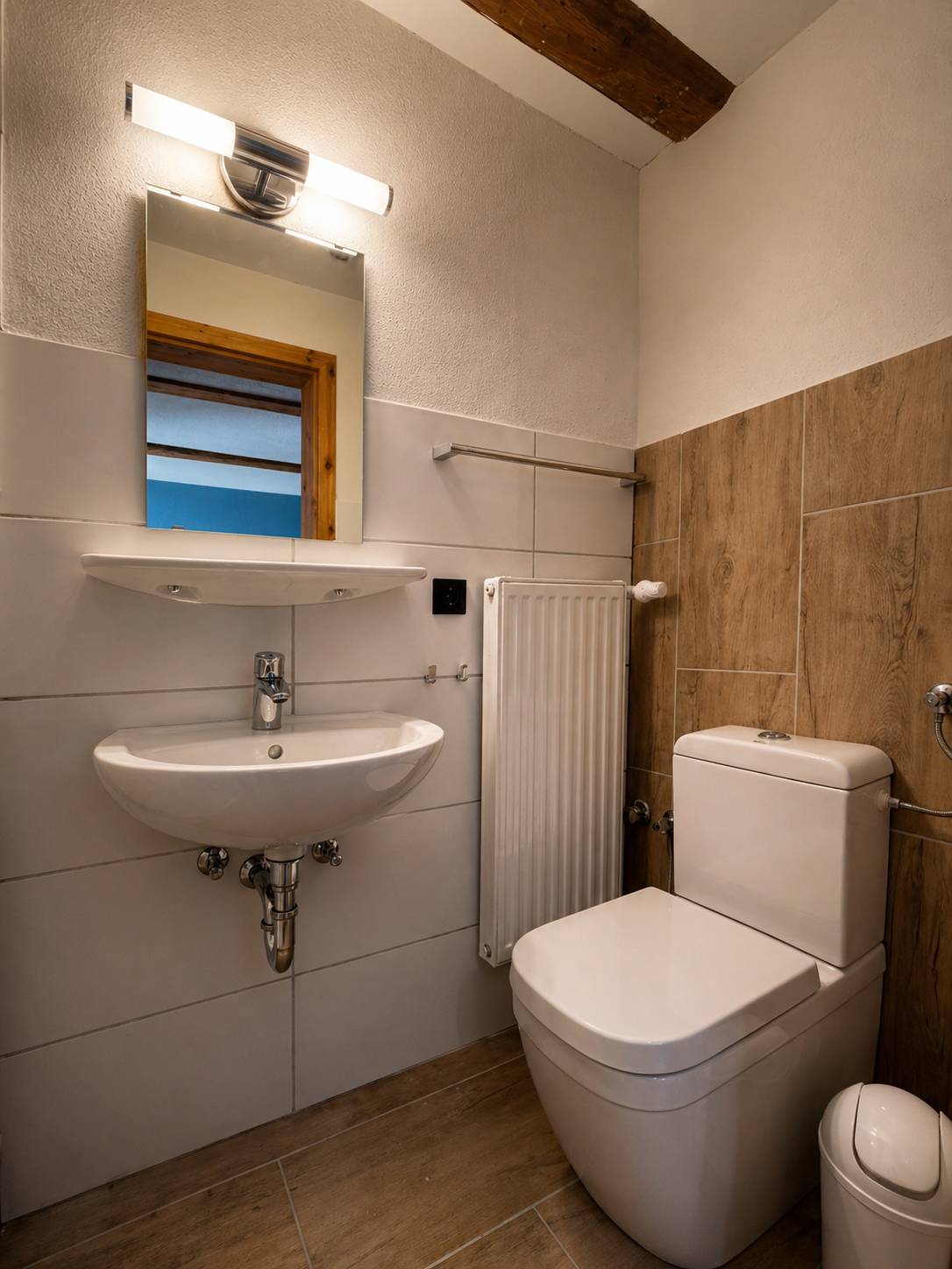 Ein kleines Badezimmer mit einem weißen Toiletten, einem Wandwaschbecken, einem Spiegel, einem Handtuchhalter, einer Heizung und einer Holz-Wandverkleidung.