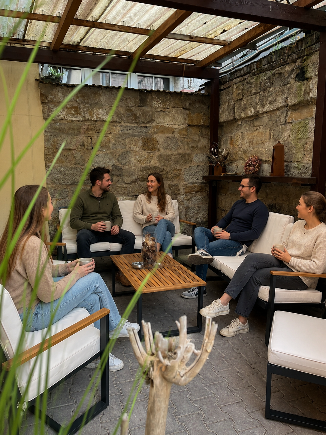 Fünf Menschen sitzen in einem Garten unter einem Dach, trinken Kaffee und unterhalten sich. Es ist eine gemütliche, steinerne Terrasse mit Holzmöbeln und Dekorationen an der Wand.