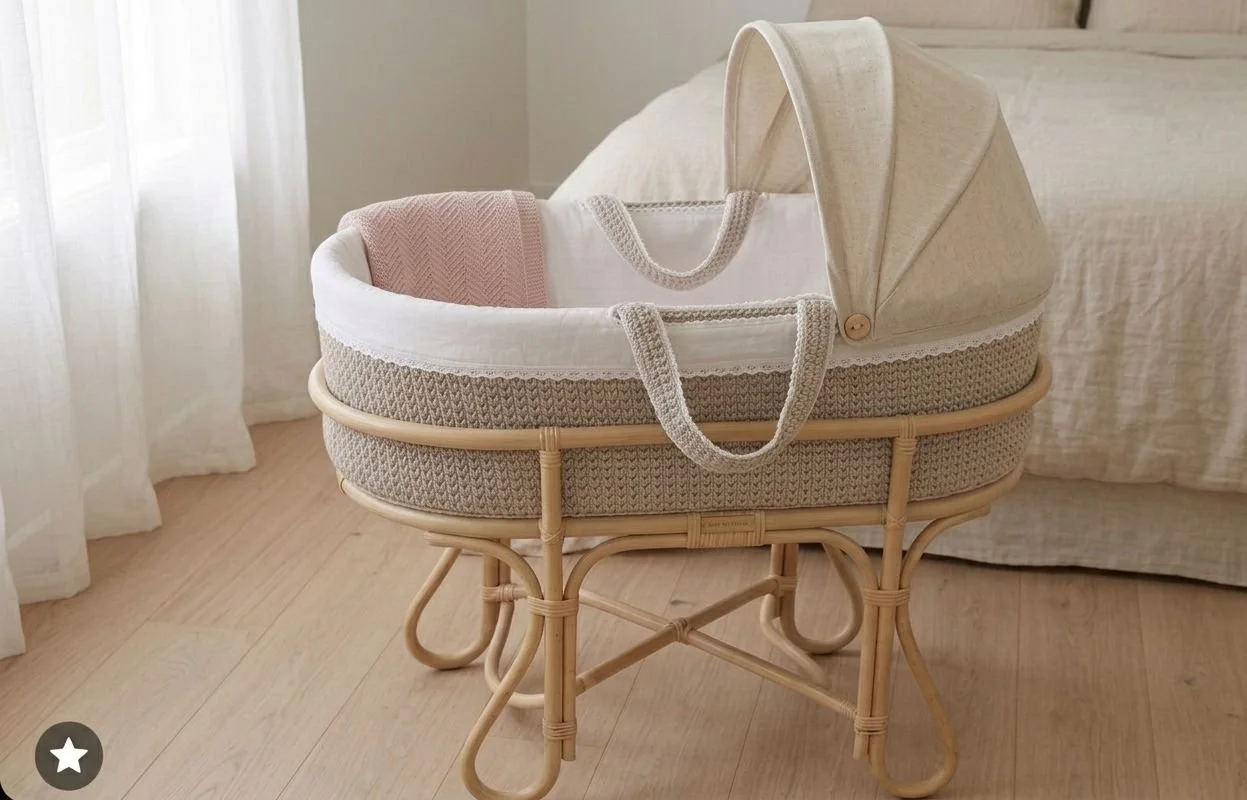 Cuna de bebé de cesto de mimbre con capota y ropa suave en interior de habitación con cortinas blancas y cama con sábanas beige.