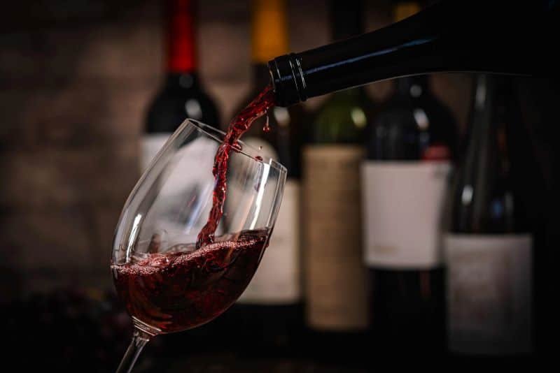 Red wine being poured into a wine glass with a number of elite bottles of wine in the background
