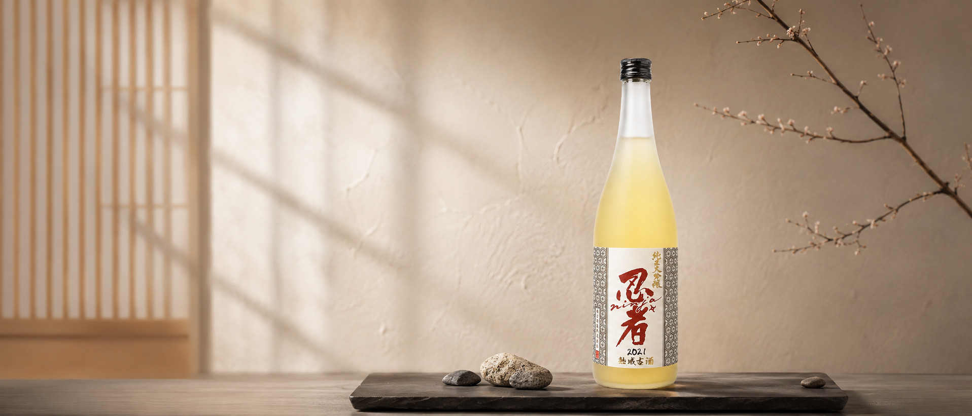 A bottle of Ninja 2021 Junmai Daiginjo Yamadanishiki Kojoon a wooden tray with three stones, set against a beige wall with a branch of blossoms on the right side and a shoji screen on the left.