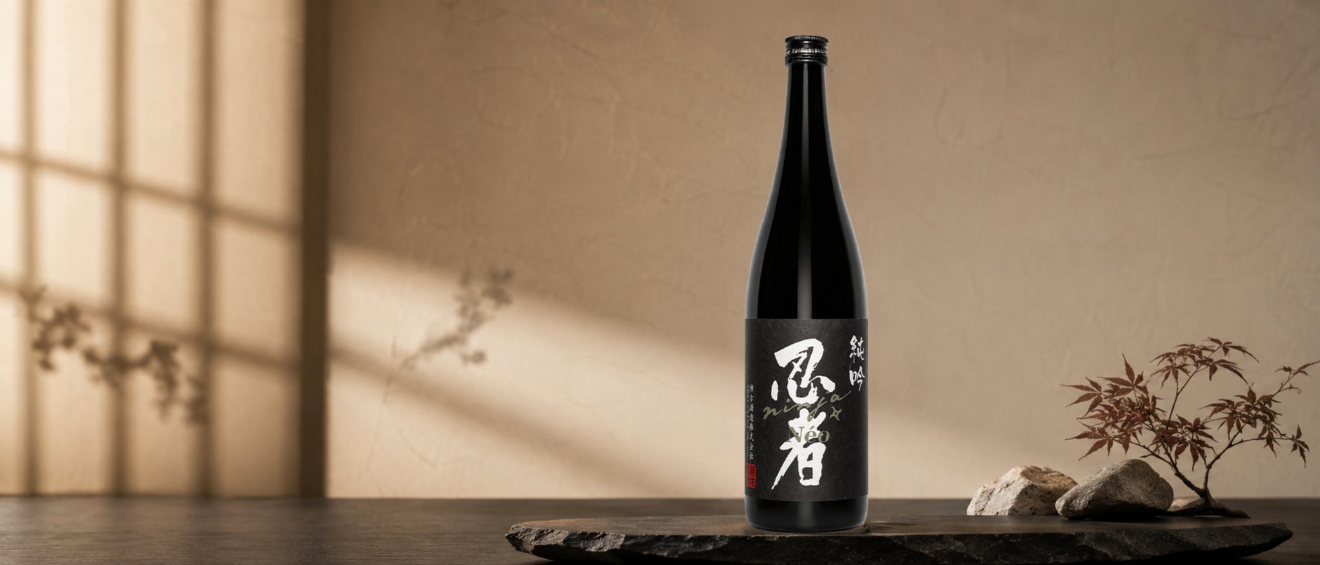 A bottle of Ninja NEO Junmai Ginjo, with Japanese characters on a black label, placed on a black surface with rocks and a small Japanese maple plant, against a beige background with soft shadow patterns.