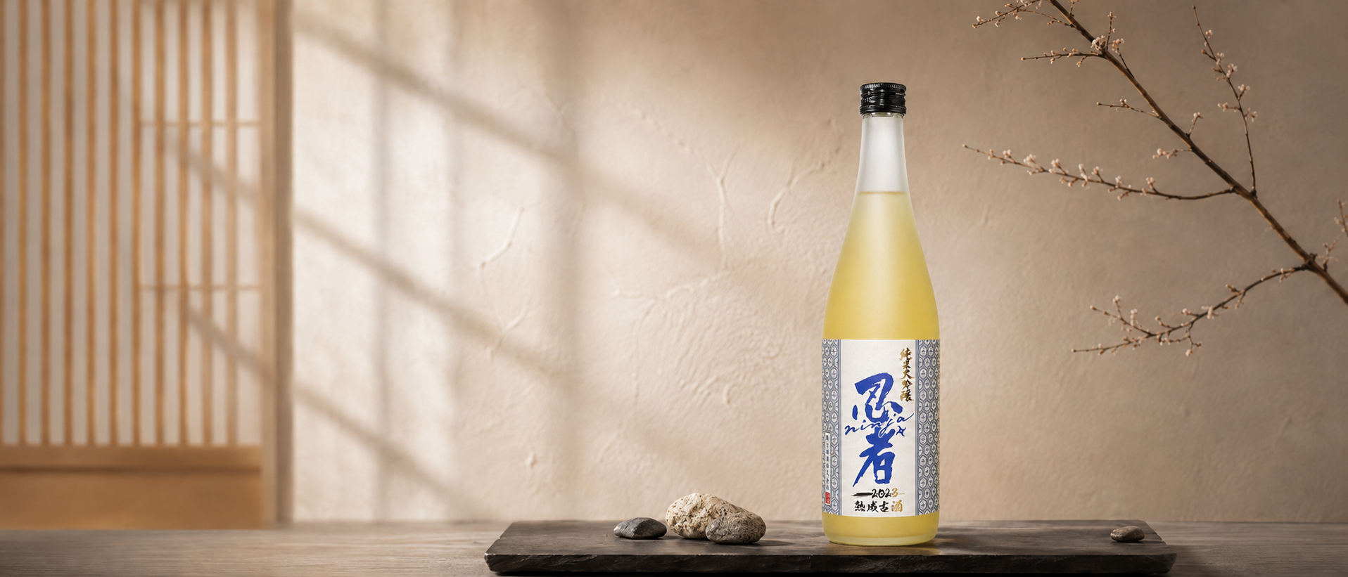 A bottle of Ninja 2023 Junmai Daiginjo Aiyama Kojo, placed on a black rectangular tray with rocks, against a beige textured wall and next to a branch with pink blossoms.