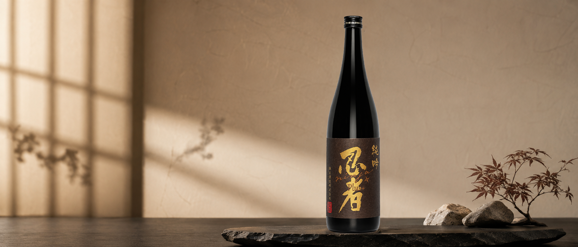 A bottle of Ninja PLUS Junmai Ginjo with Japanese characters, placed on a black wooden surface, with rocks and a small Japanese maple branch nearby, against a beige background with soft shadowed lighting.