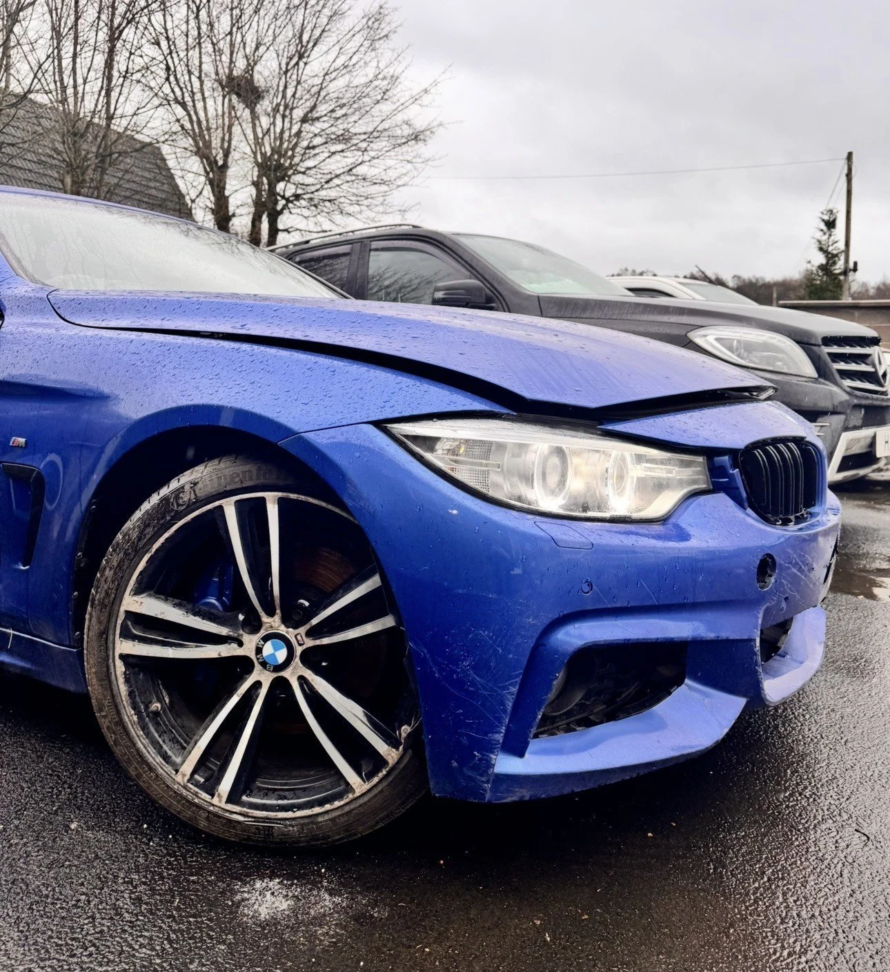 Blue BMW sedan with front-end damage parked on wet pavement, with other cars and leafless trees in the background.