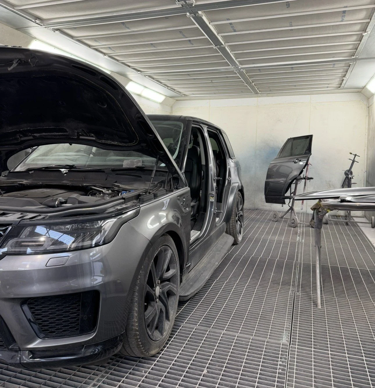 An automotive repair garage with a silver SUV having its front hood open, a door removed from its frame, and other car parts on stands in the background.