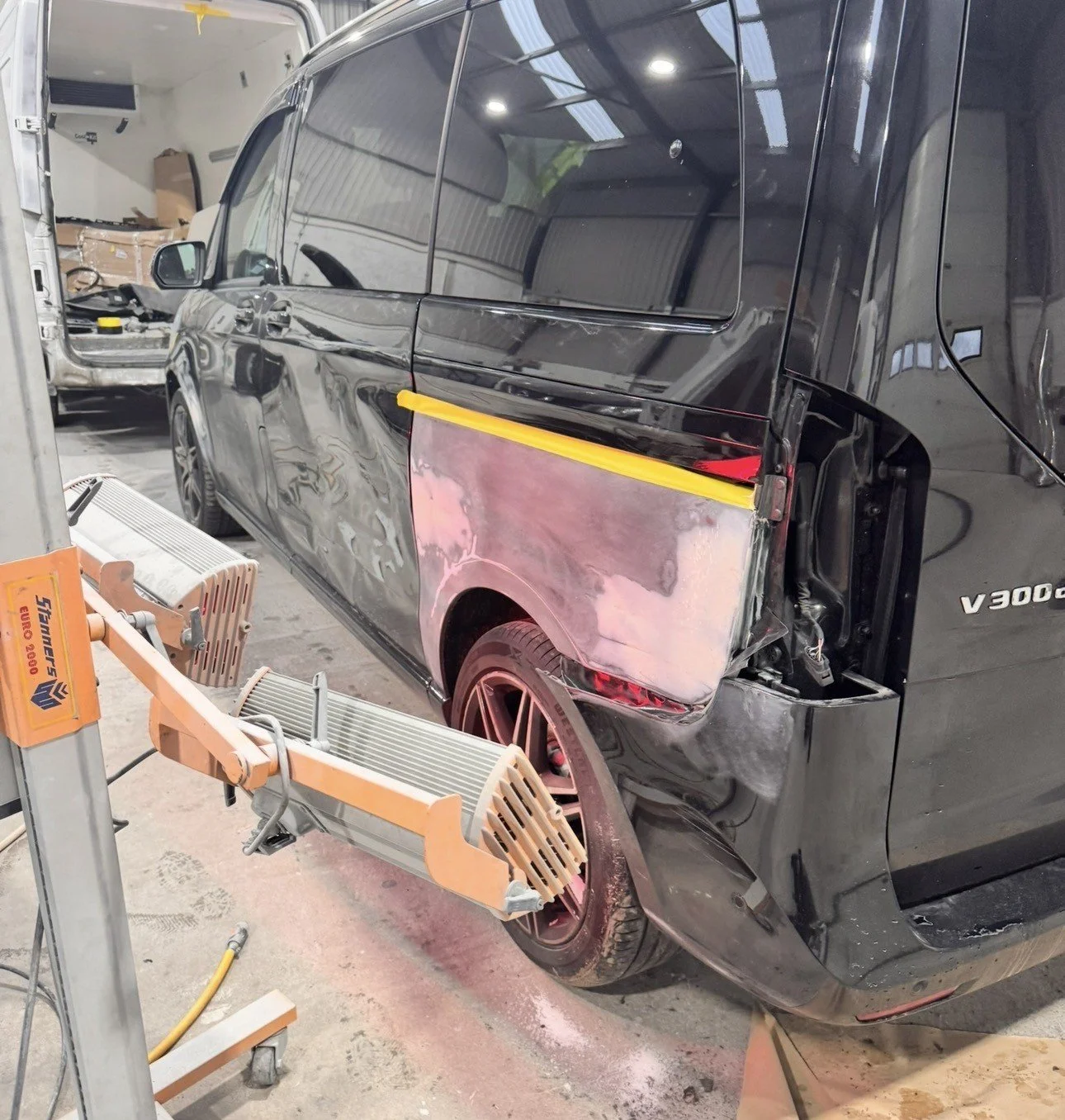 Black Mercedes-Benz van being repaired in a garage, with damage on the side door and primer application on part of the body.