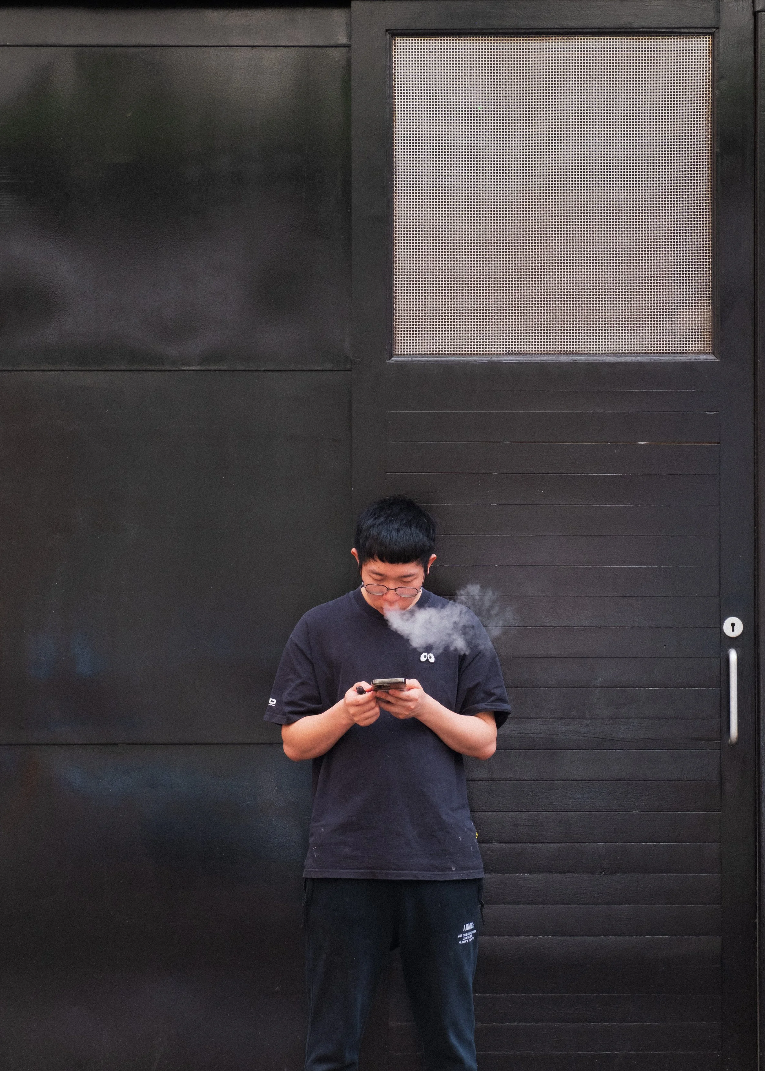 Man in all black smokes a vape while checking his phone