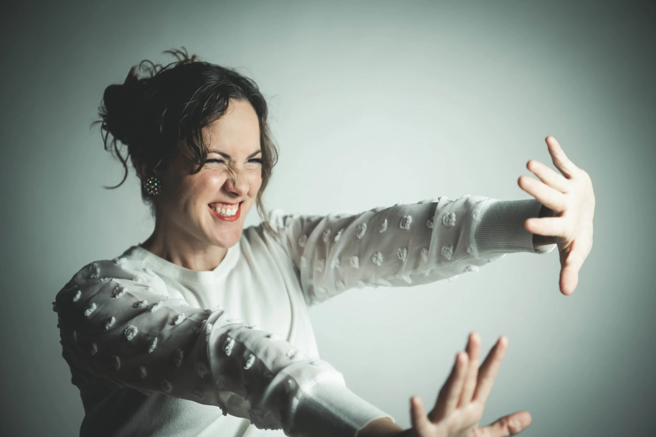 A woman, Aimee Bobruk, smiling broadly and making a playful gesture with her hands, wearing a white sweater with textured polka dots, against a plain light-colored background.