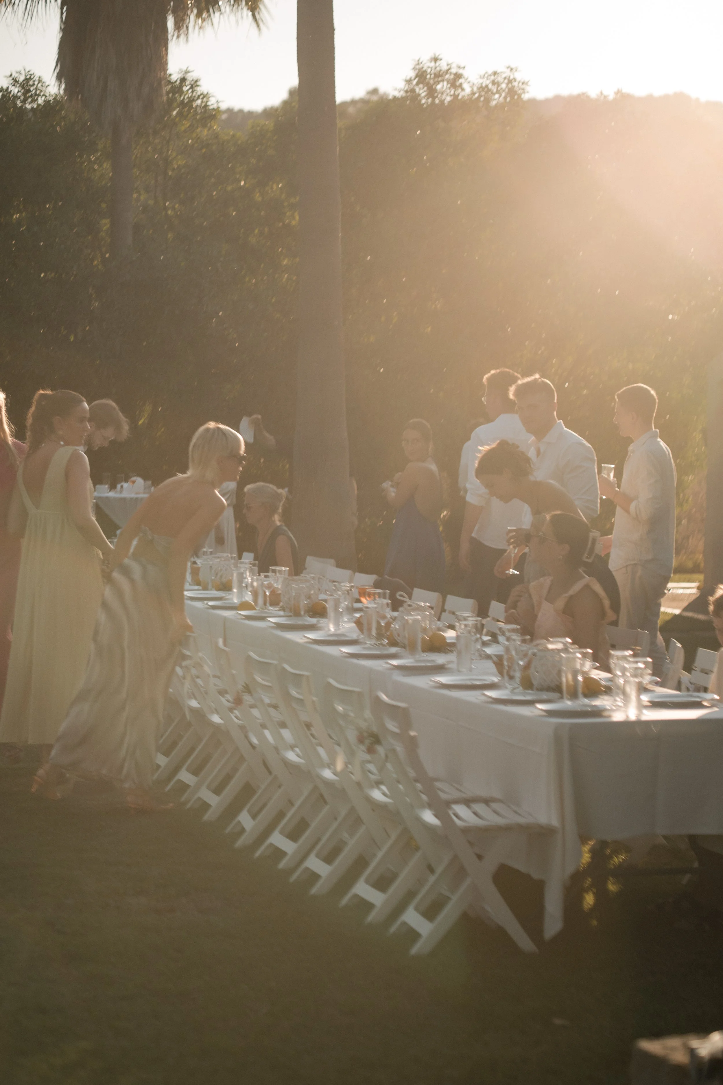 Outdoor banquet setup with a long white table, white chairs, and a group of people dressed in semi-formal attire, with trees and sunlight in the background.