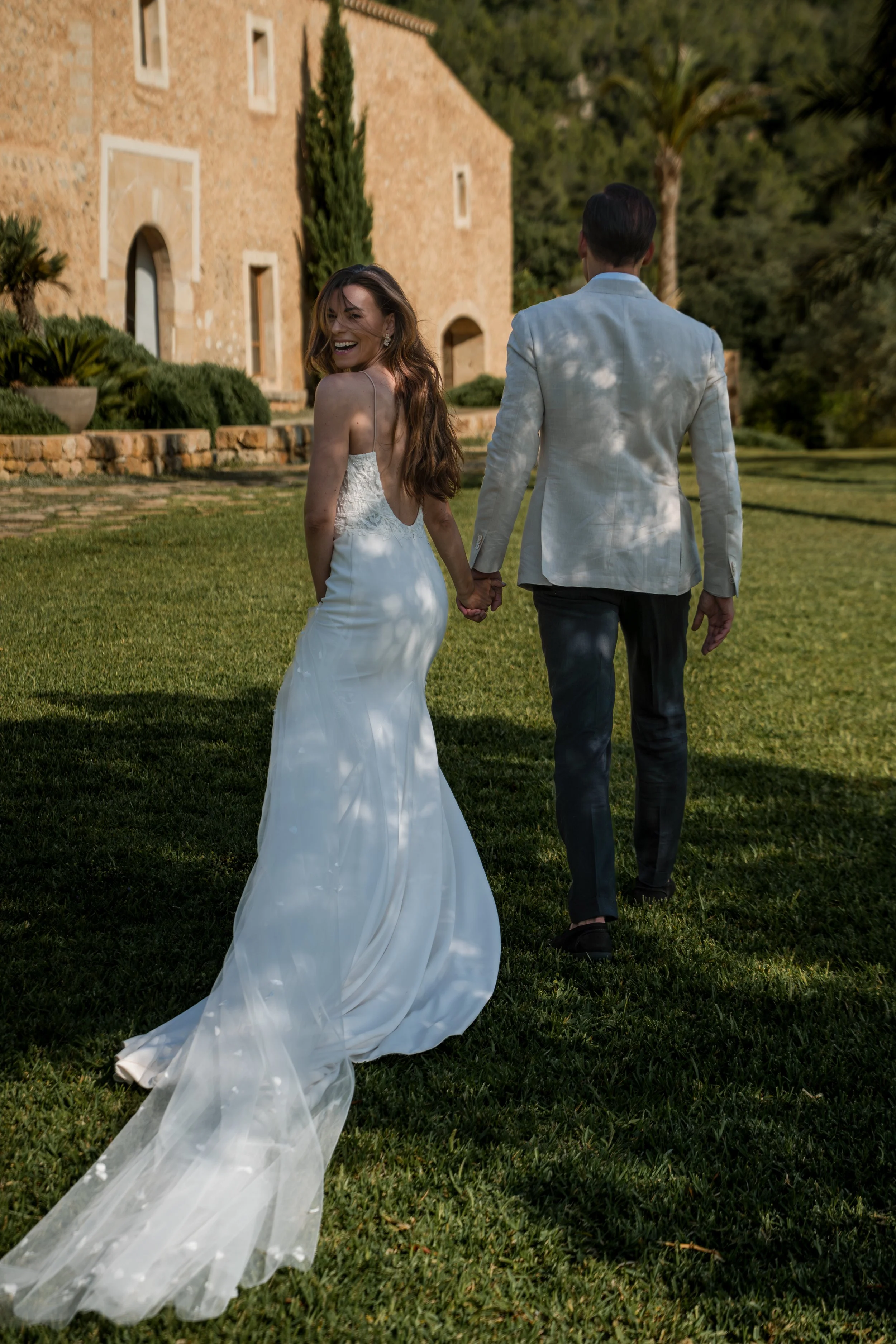 A happy woman in a white wedding dress holding hands with a man in a light gray suit, walking on a lush green lawn in front of a stone building, surrounded by trees, during sunset.